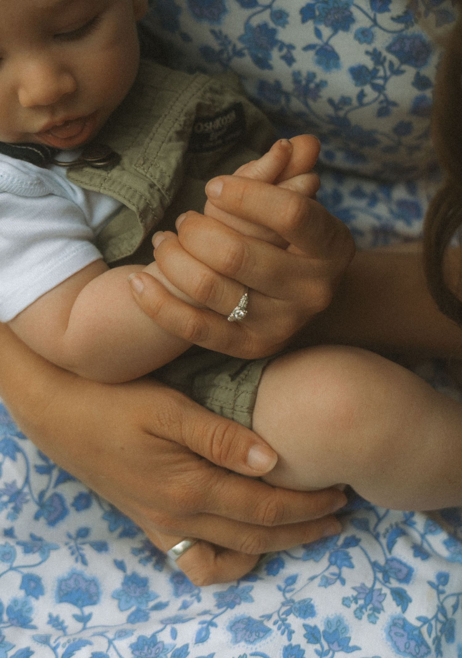 A mother gently holds her sleeping baby while wearing a diamond engagement ring, sitting on a floral blue blanket.