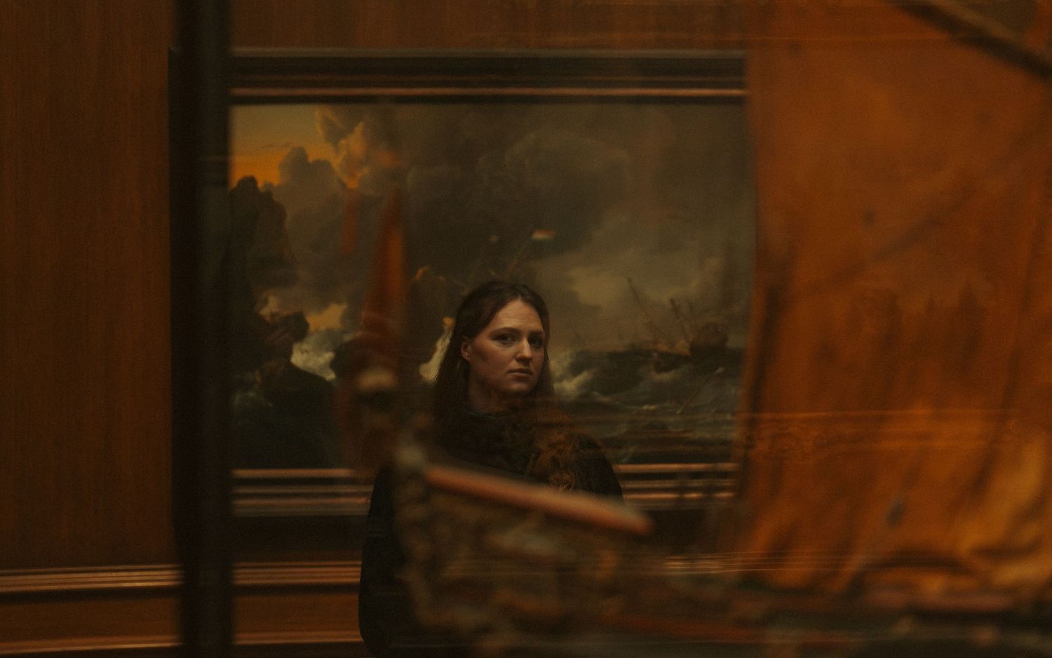 A dramatic scene from a dark movie shows someone pointing a gun while standing in a dimly lit room with paintings.