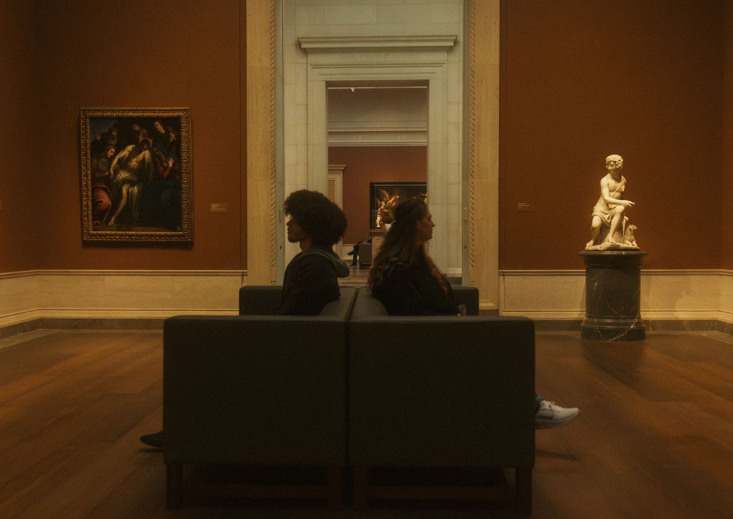 Visitors sit on a bench while viewing artwork in a dimly lit museum gallery space.