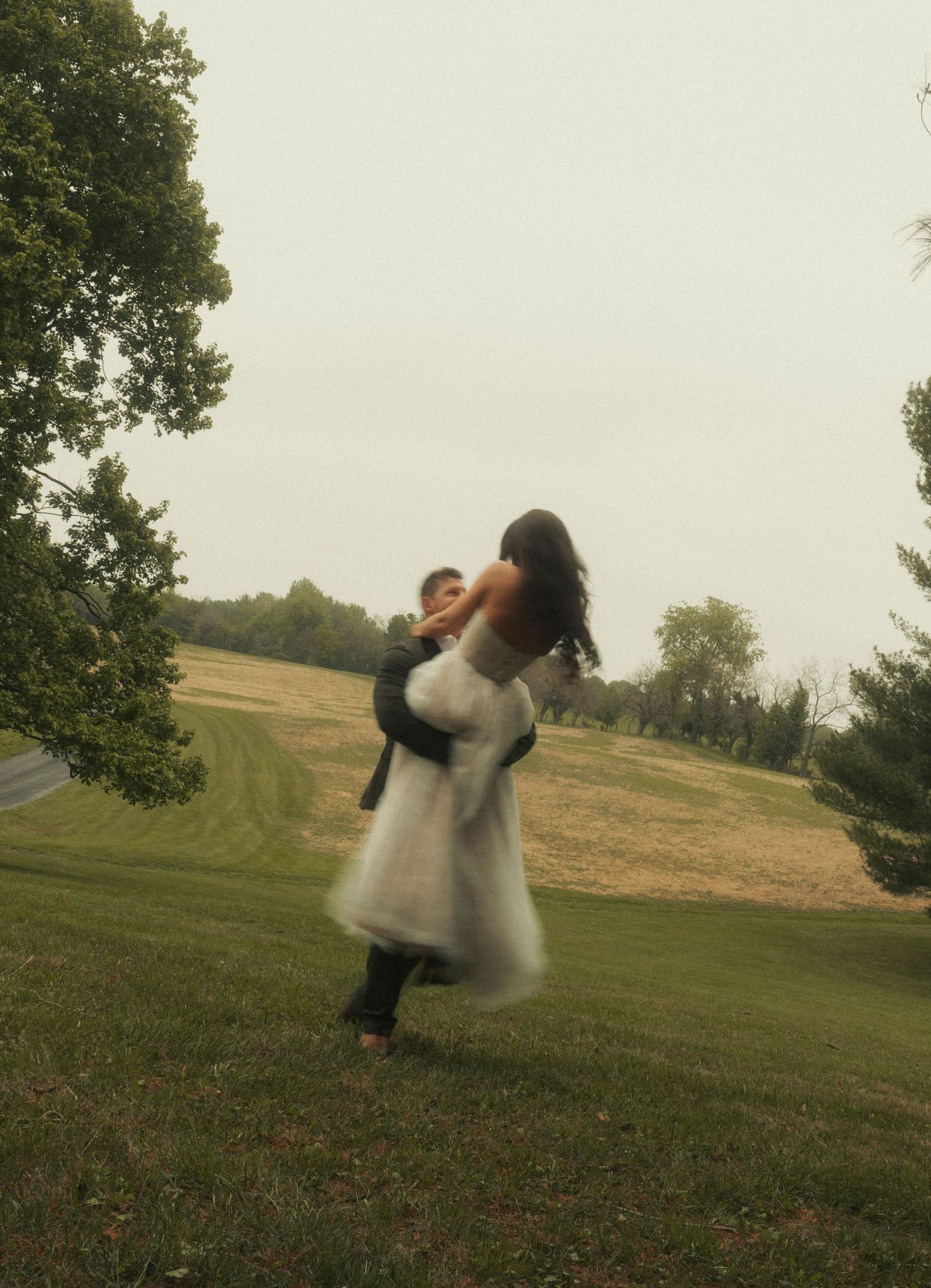 A bride in a white gown joyfully twirls with a child in her arms on a lush green lawn.