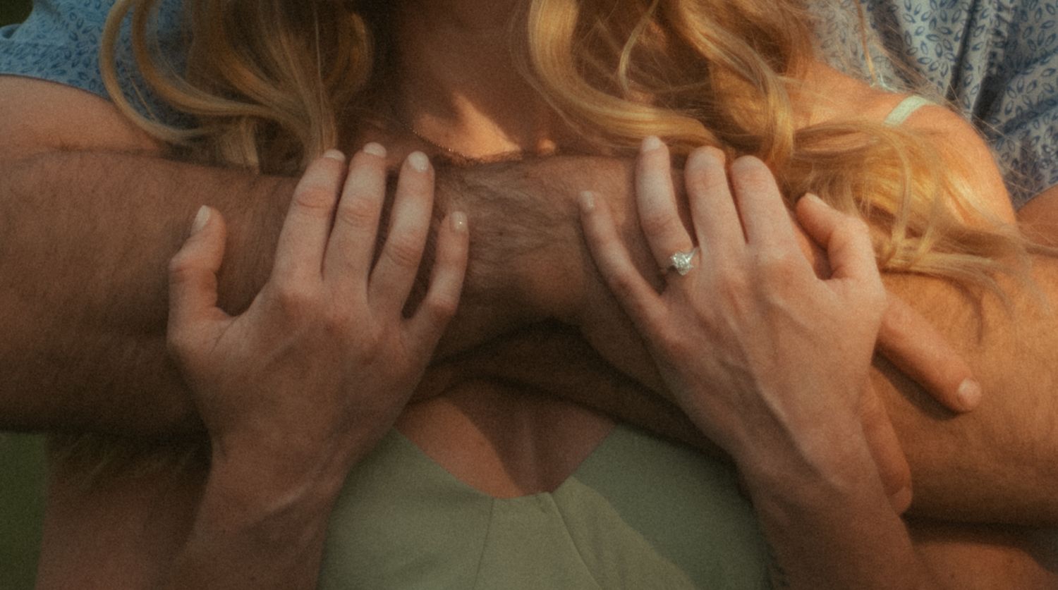 Close-up of a woman in a green top with a man's hands resting on her shoulders, she wears a diamond ring.