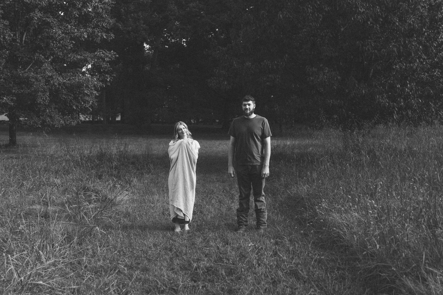 Black and white photo of a couple standing in a grassy field surrounded by trees.