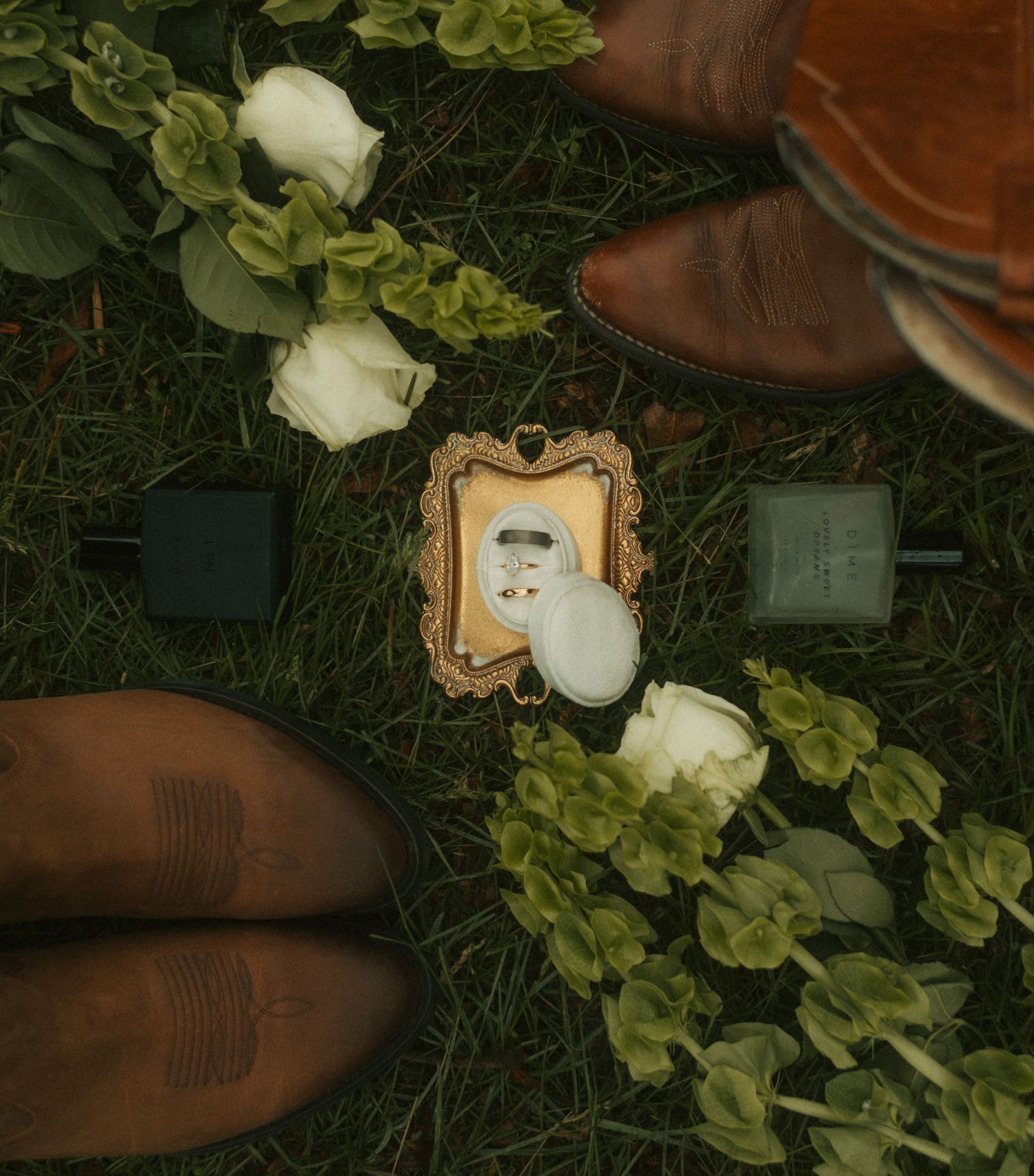 Two people in cowboy boots stand over white roses, greenery, a framed photo, and nail polish on grass.