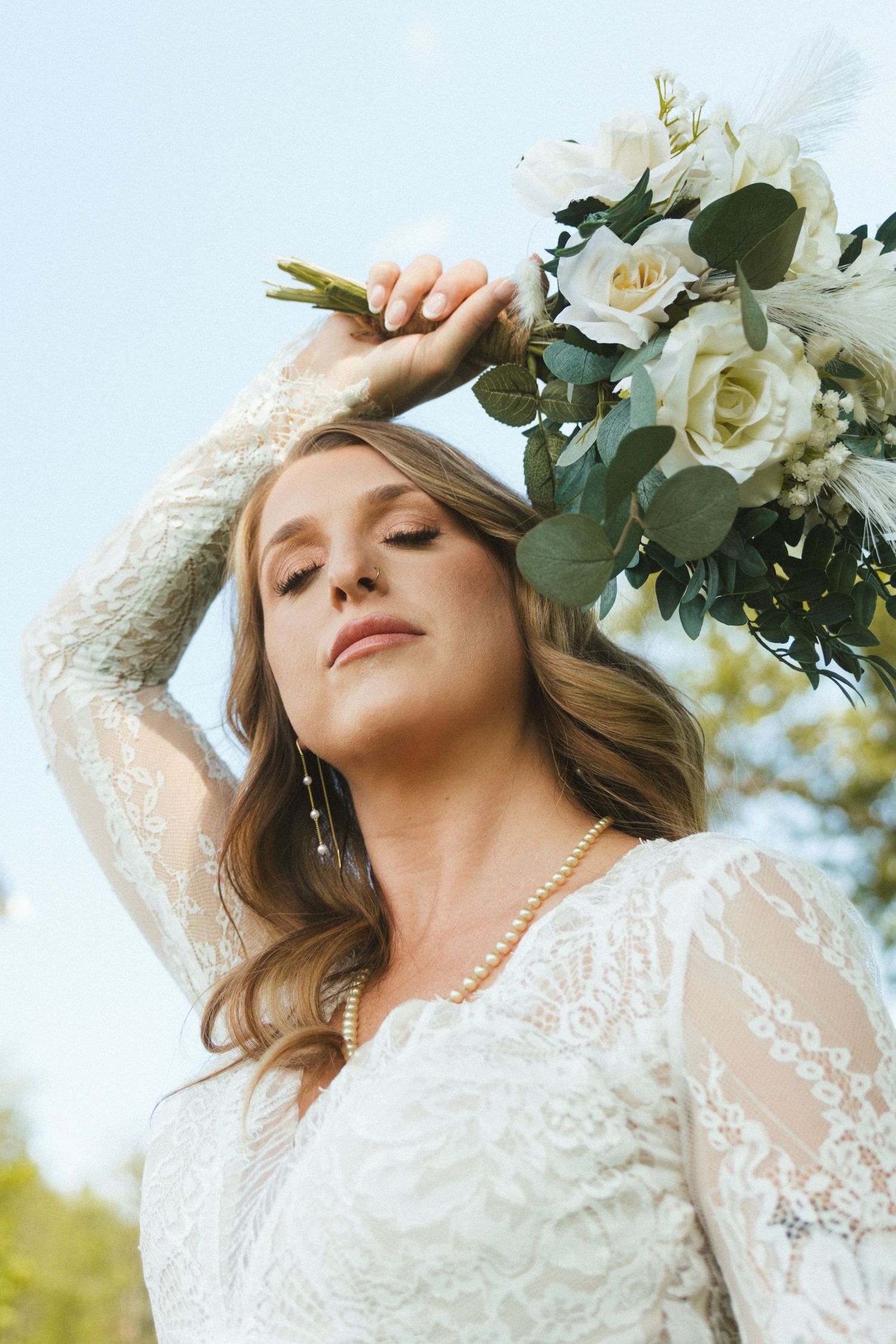 Bride in lace gown holds white floral bouquet overhead with eyes closed outdoors.