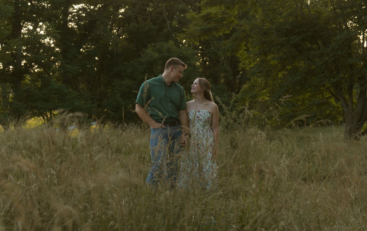 A couple stands together in a golden meadow surrounded by lush green trees during a warm sunset.