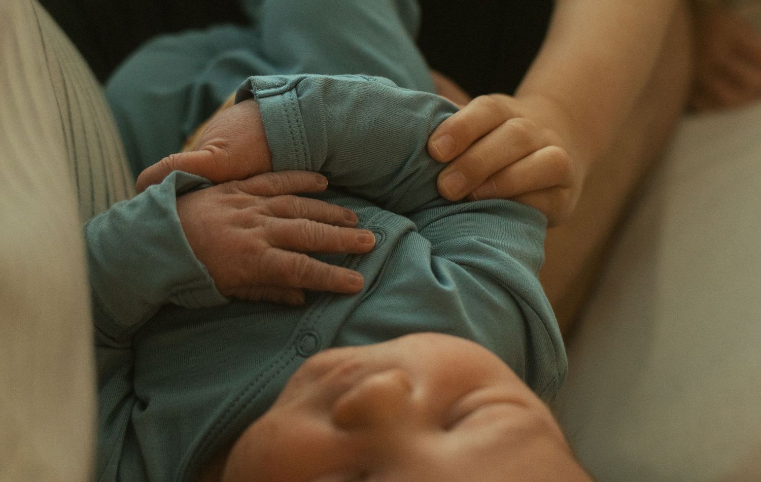 Close-up of a newborn baby in teal onesie cradled in adult arms, tiny hands visible.