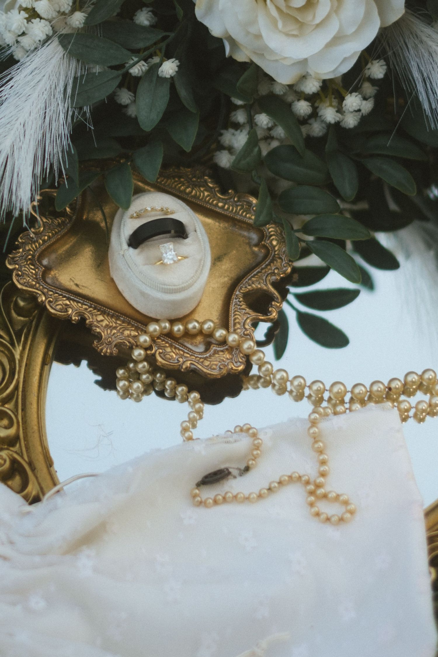 Elegant wedding rings in gold velvet box with pearl necklace, white roses, and greenery on marble surface.