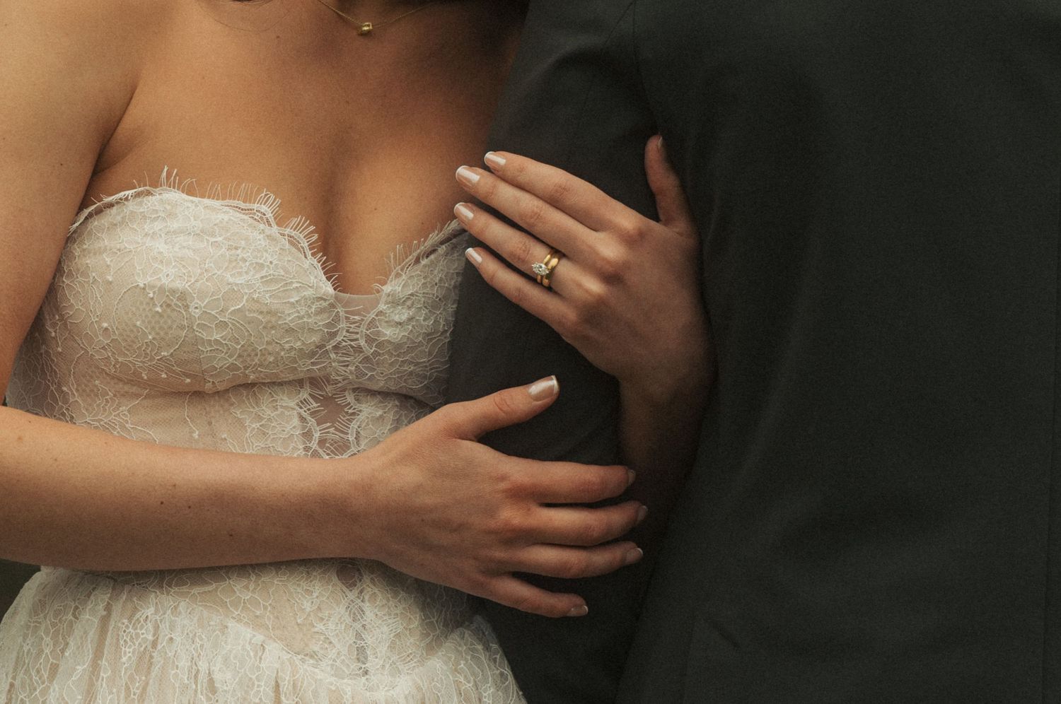 Bride in lace wedding gown embraced by groom in dark suit, showcasing diamond engagement ring on her hand.