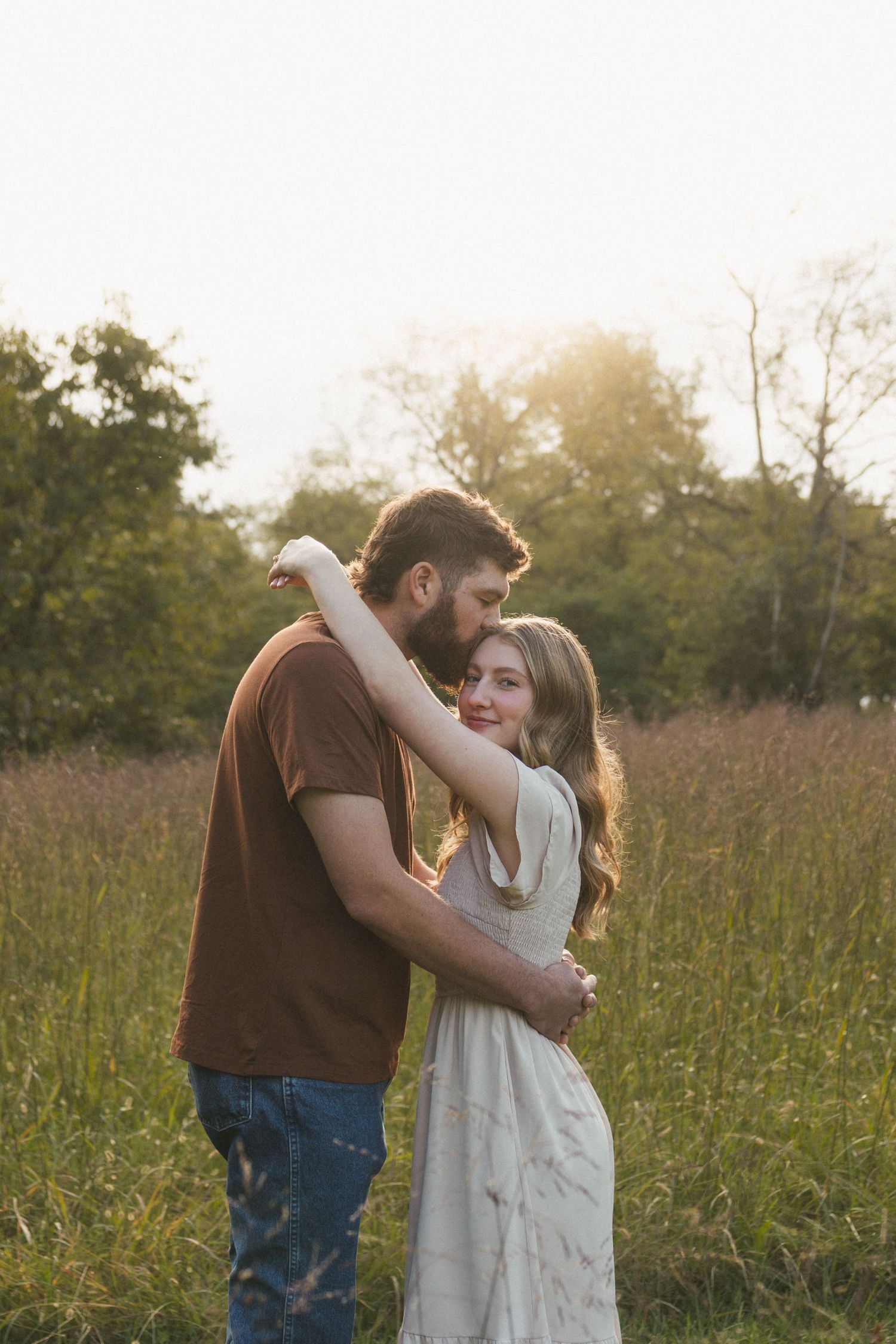 A couple embraces in a sunlit meadow, man in brown shirt kissing woman's head as she smiles softly.