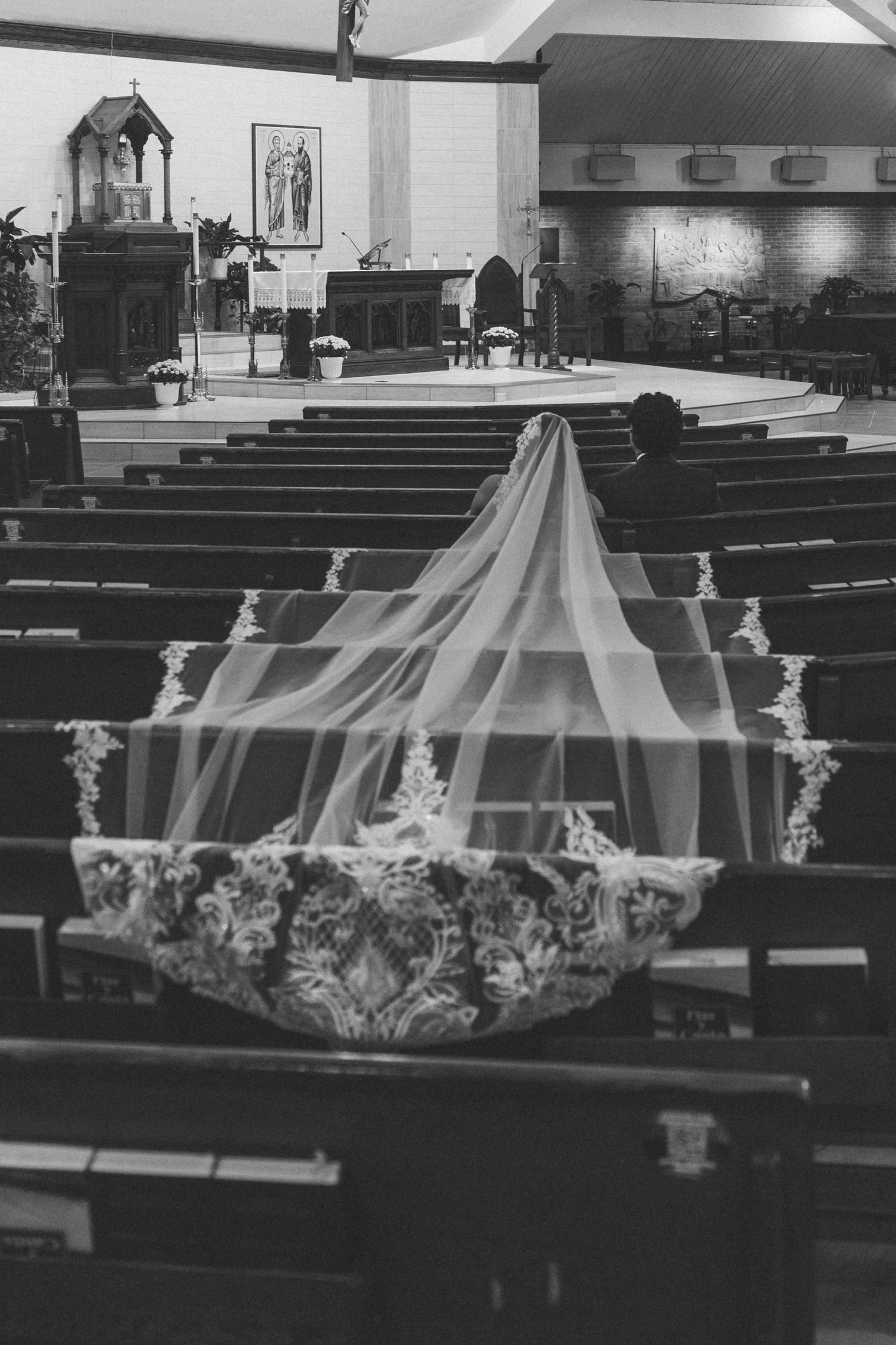 Black and white photo of a bride in a long veil sitting alone in church pews before her wedding ceremony.