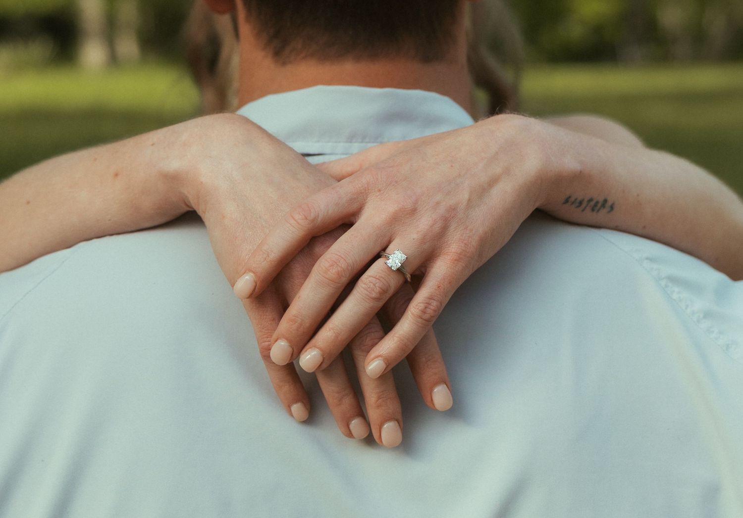 Close-up of woman\u2019s hand with engagement ring resting on man\u2019s shoulder outdoors.