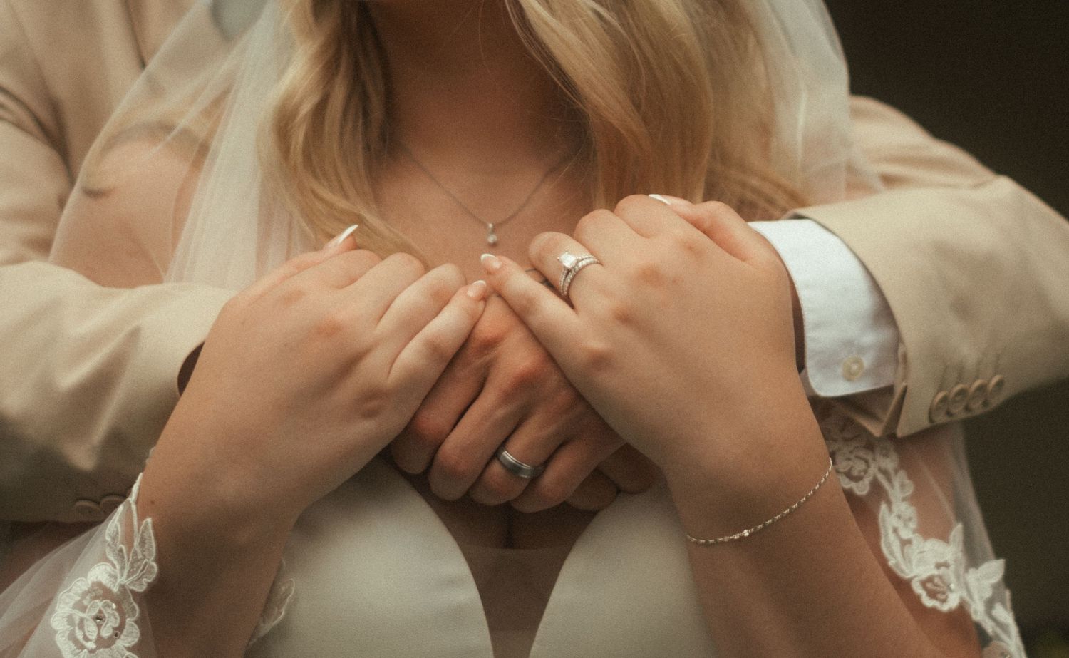 Bride and groom holding hands showing wedding rings, bride wearing lace dress and veil with delicate necklace.