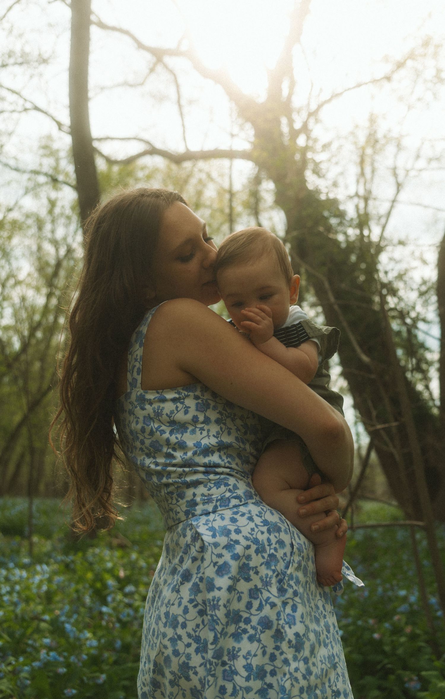 A mother in a floral dress tenderly kisses her baby while standing in a sunlit forest with blue wildflowers.