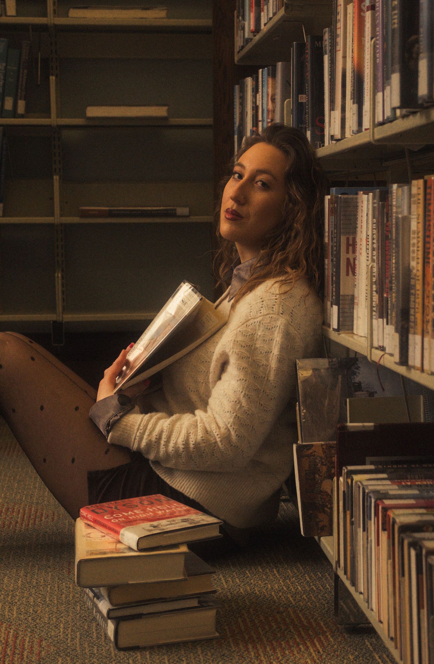 A person sits on the floor of a library reading a book surrounded by bookshelves in warm lighting.