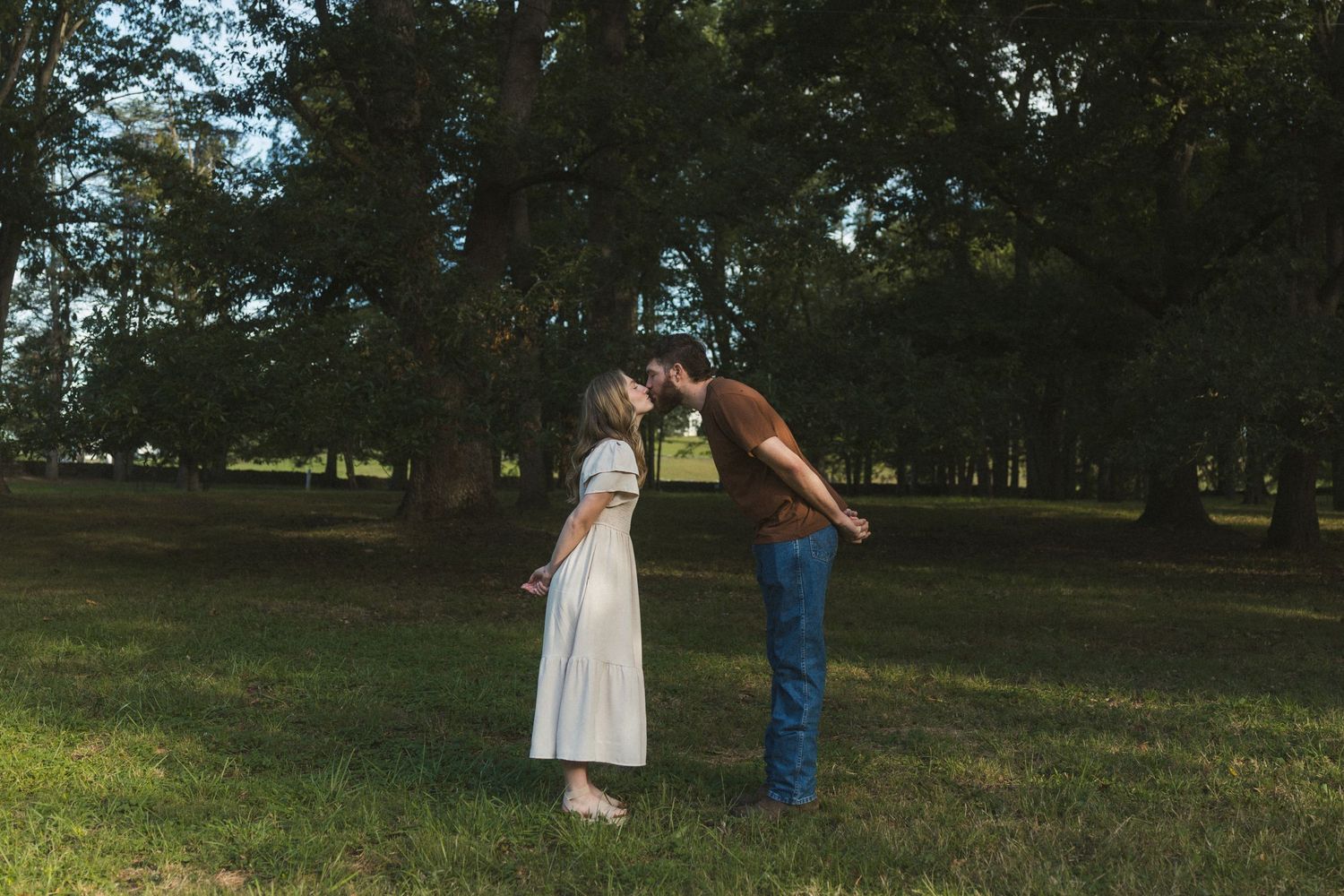 Couple sharing a romantic kiss in a lush green park surrounded by tall trees on a warm sunny day.