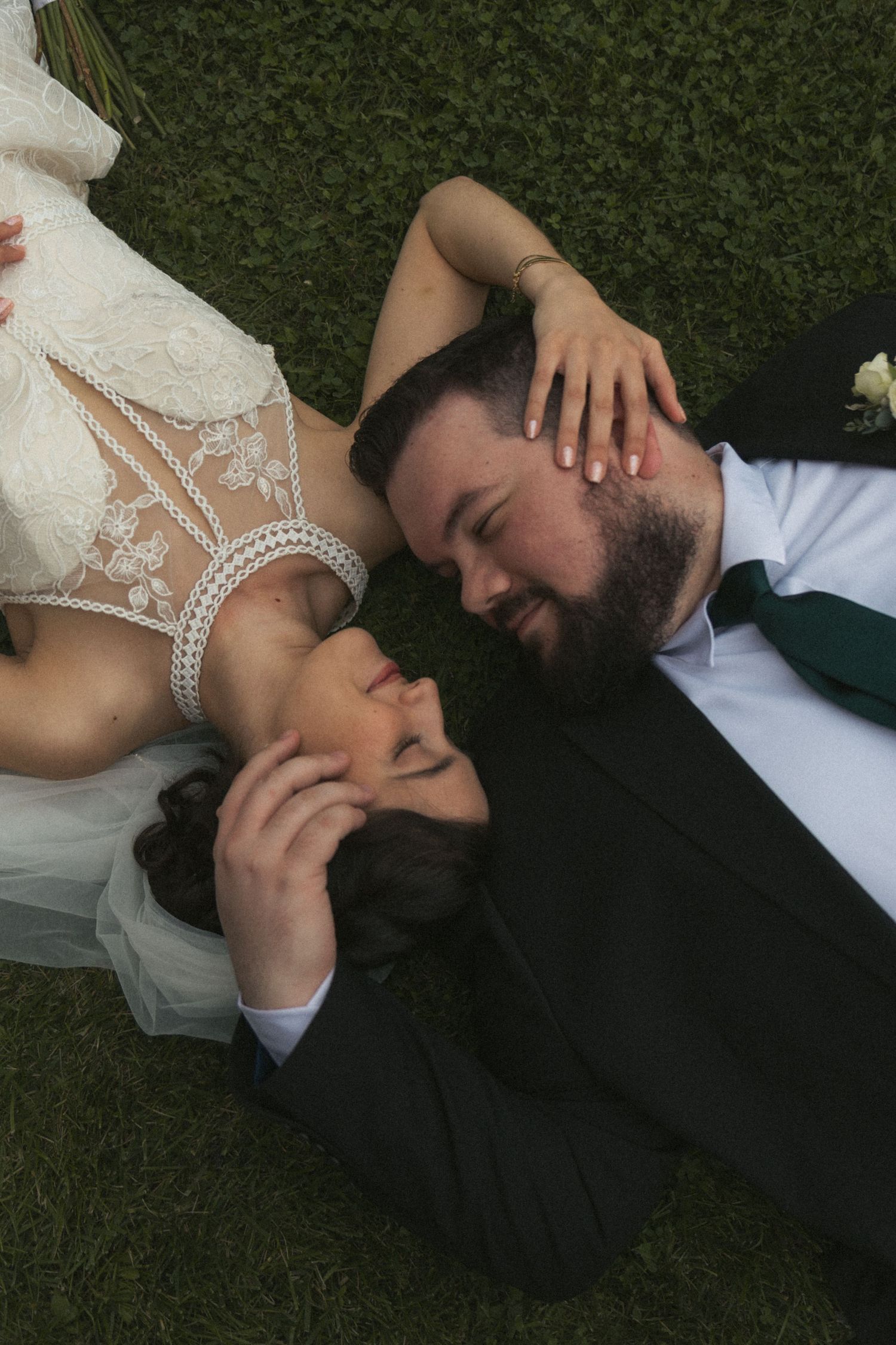 Bride and groom lying on grass, sharing a tender moment in wedding attire, with the bride in lace dress and groom in dark suit.