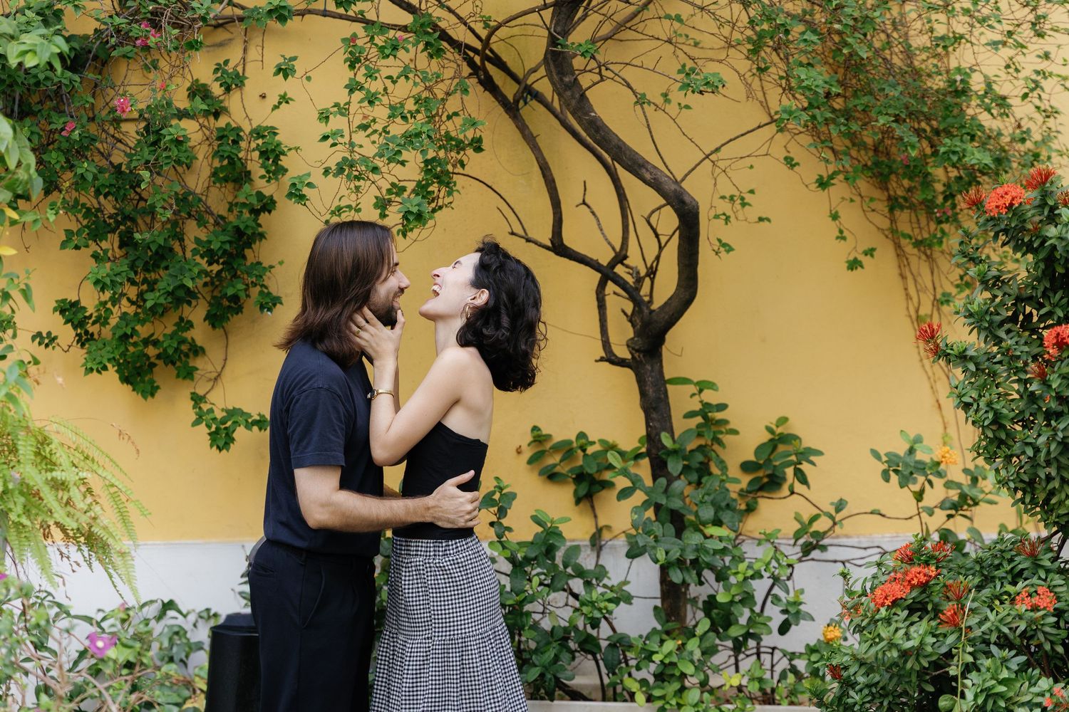 A romantic couple embraces beneath a flowering vine on a yellow wall in a garden setting.