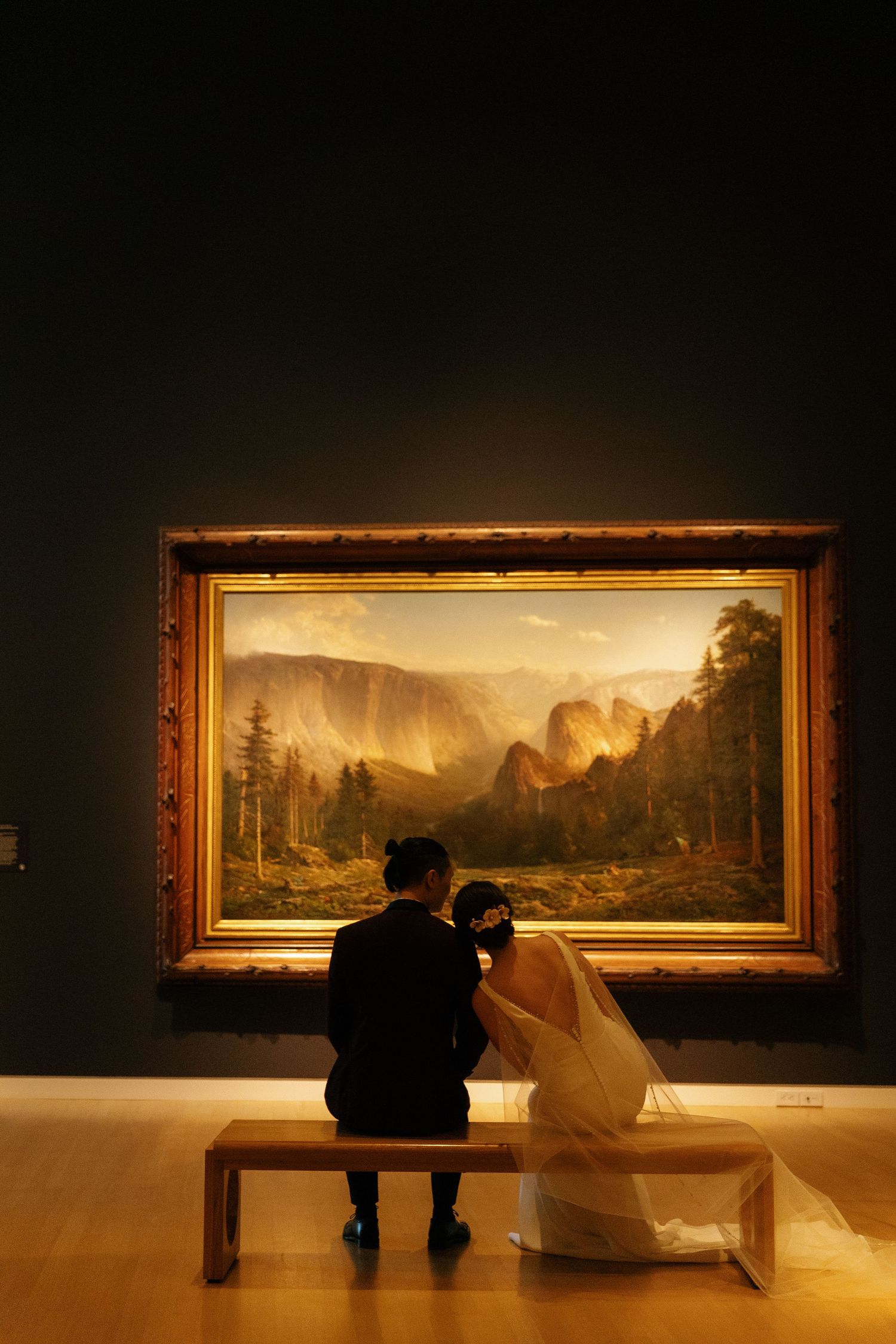 Newlyweds sitting on bench admiring large landscape painting in dimly lit art museum.