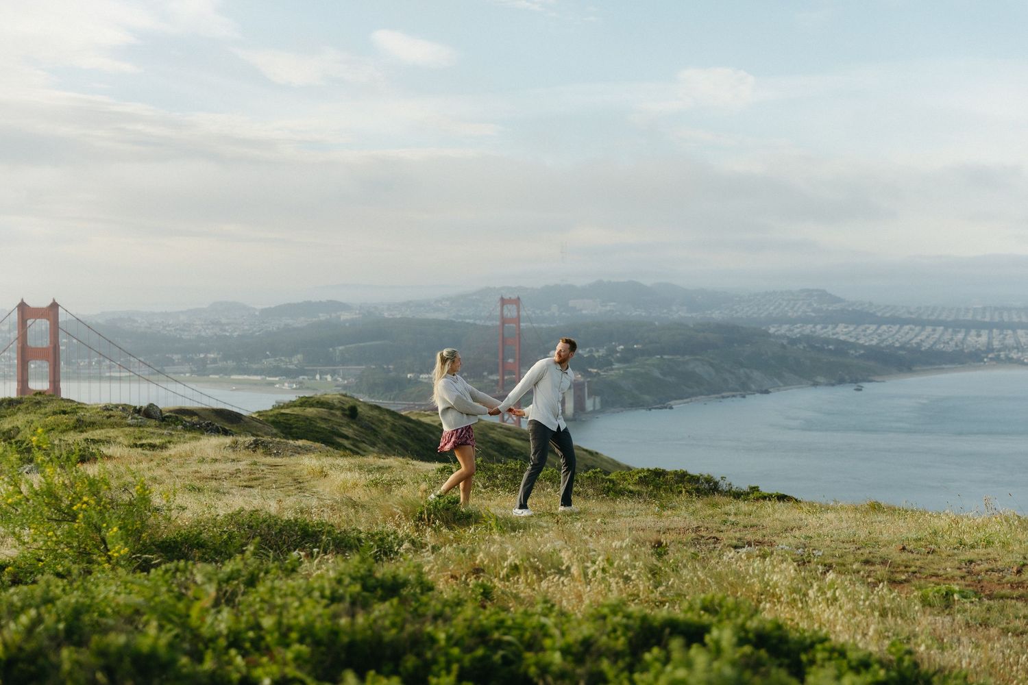 Couple holding hands running on grassy hillside with Golden Gate Bridge and bay in background.