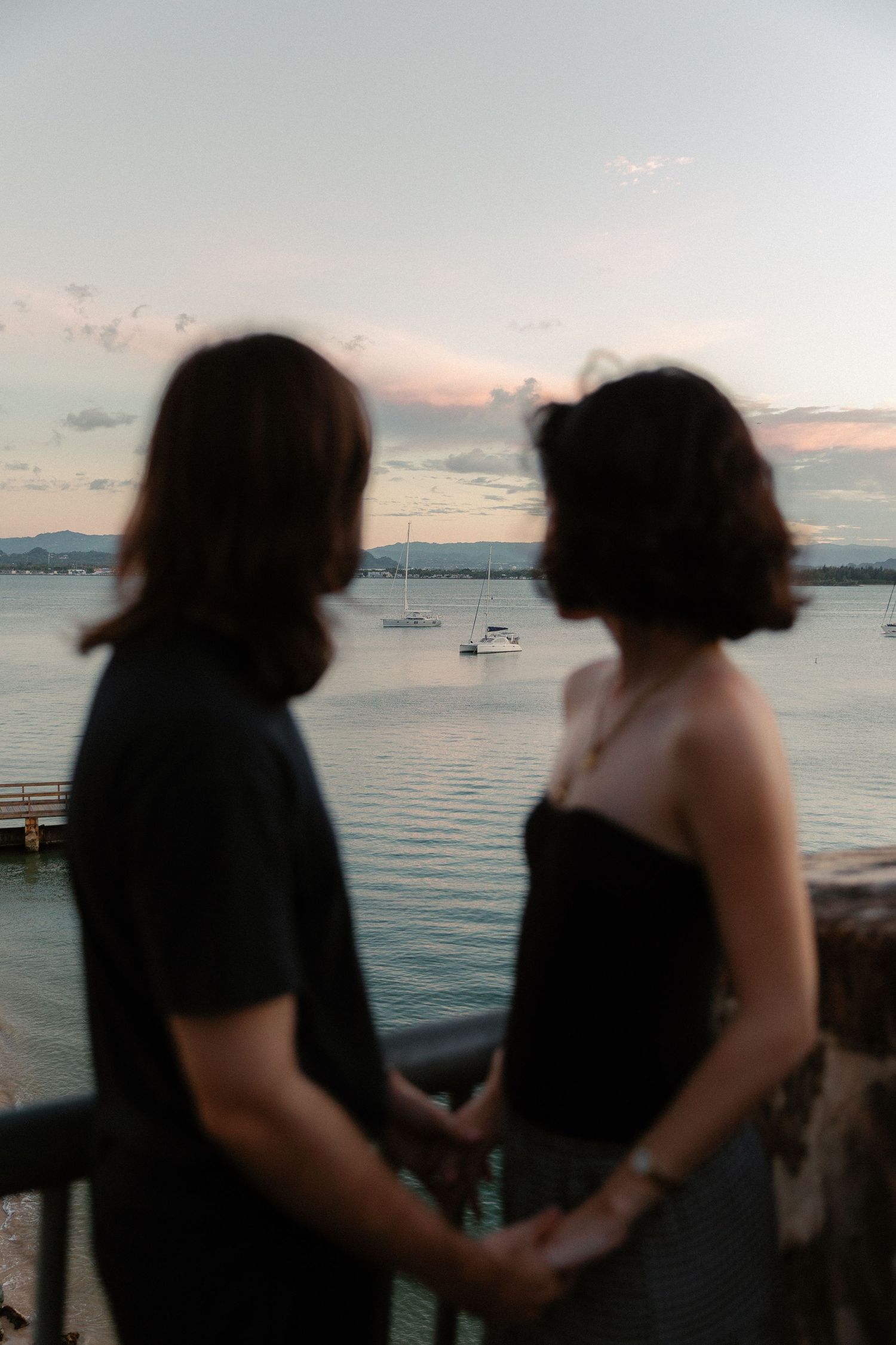 Two silhouettes stand at a waterfront railing holding hands while watching boats on calm water at sunset.