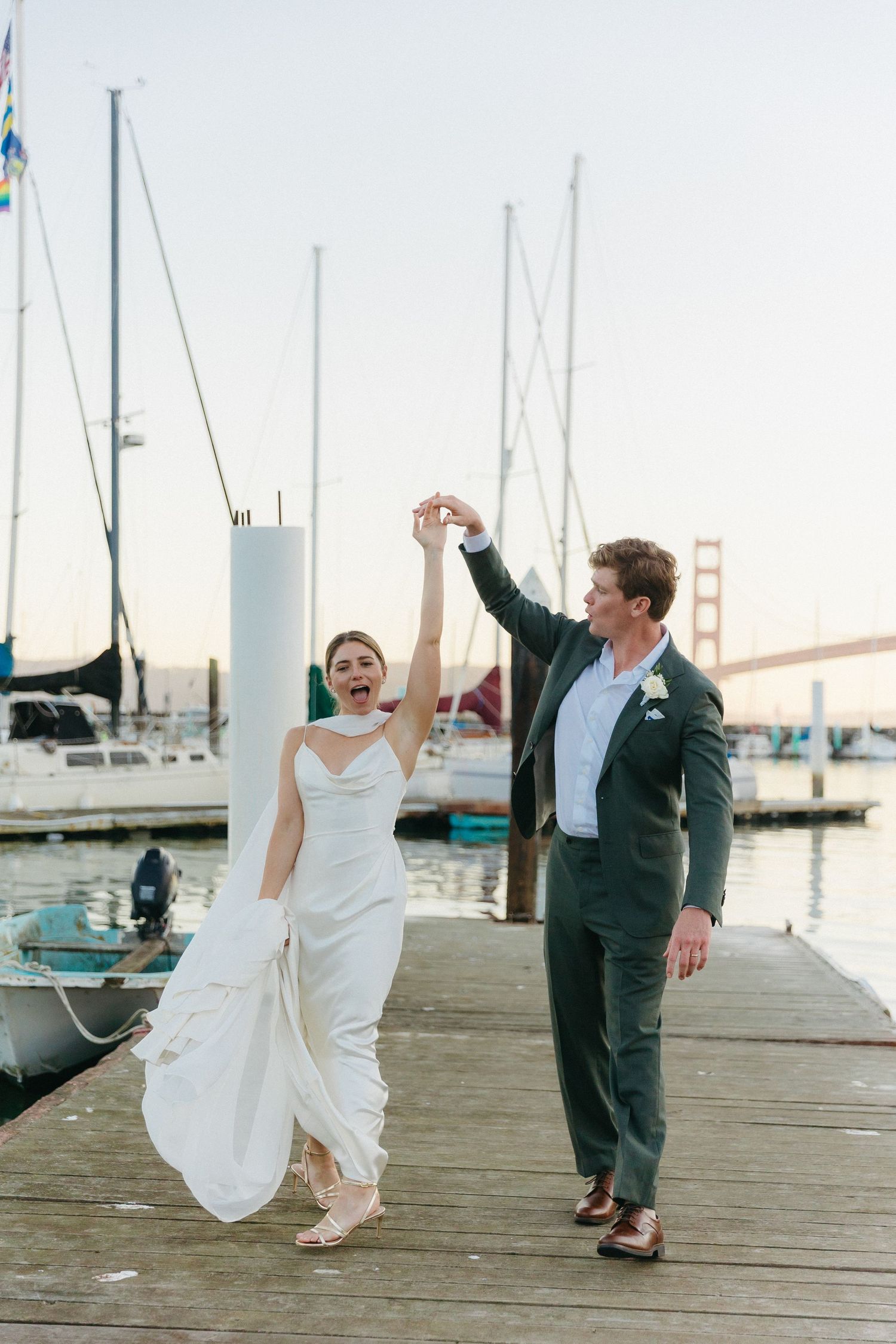 Joyful bride being twirled by groom on a marina dock with Golden Gate Bridge in background.