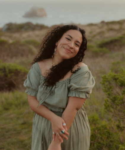 Young woman with curly hair smiling in sage green dress against coastal greenery at dusk.