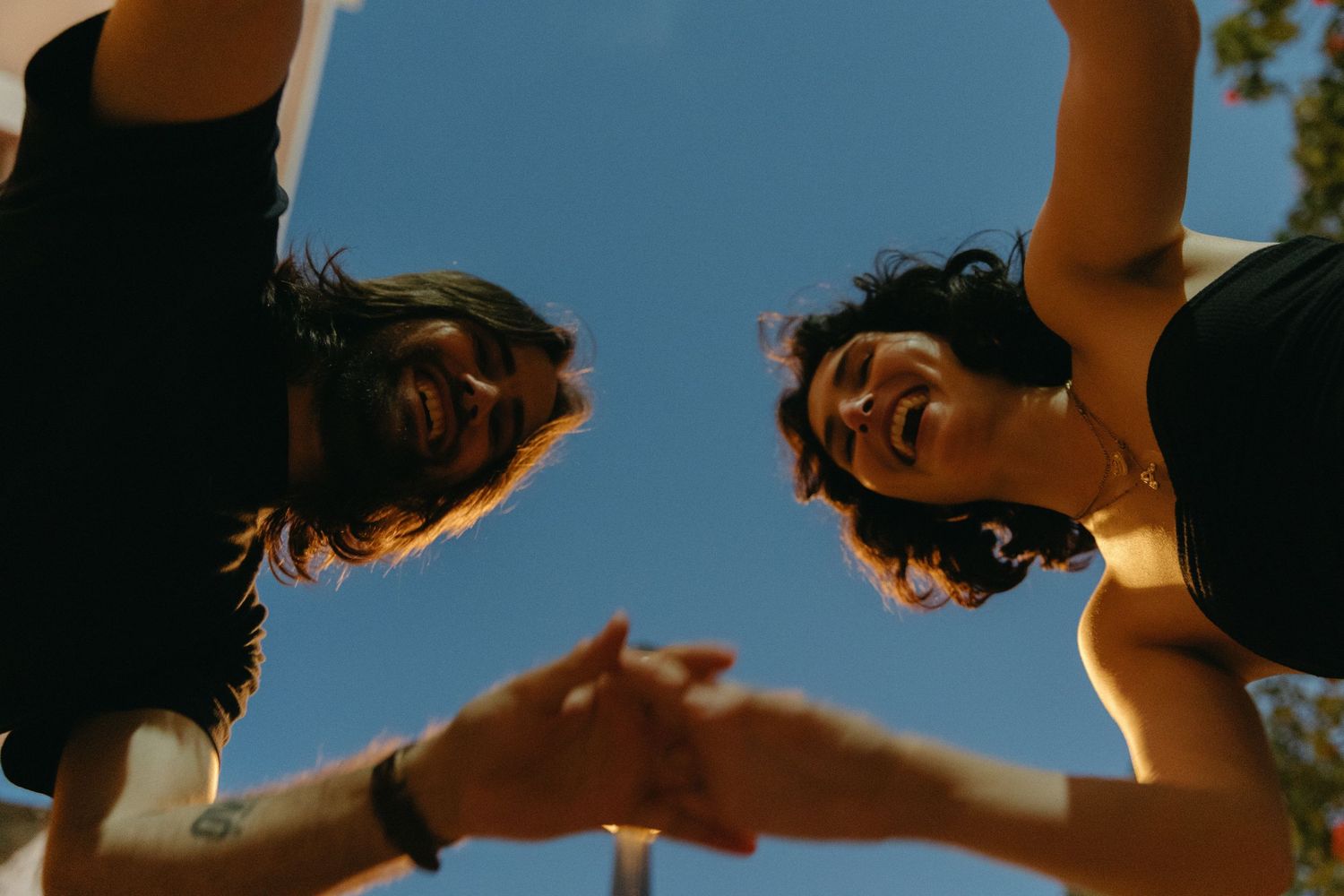 Two people lean in close from above and reach toward each other against a bright blue sky background.