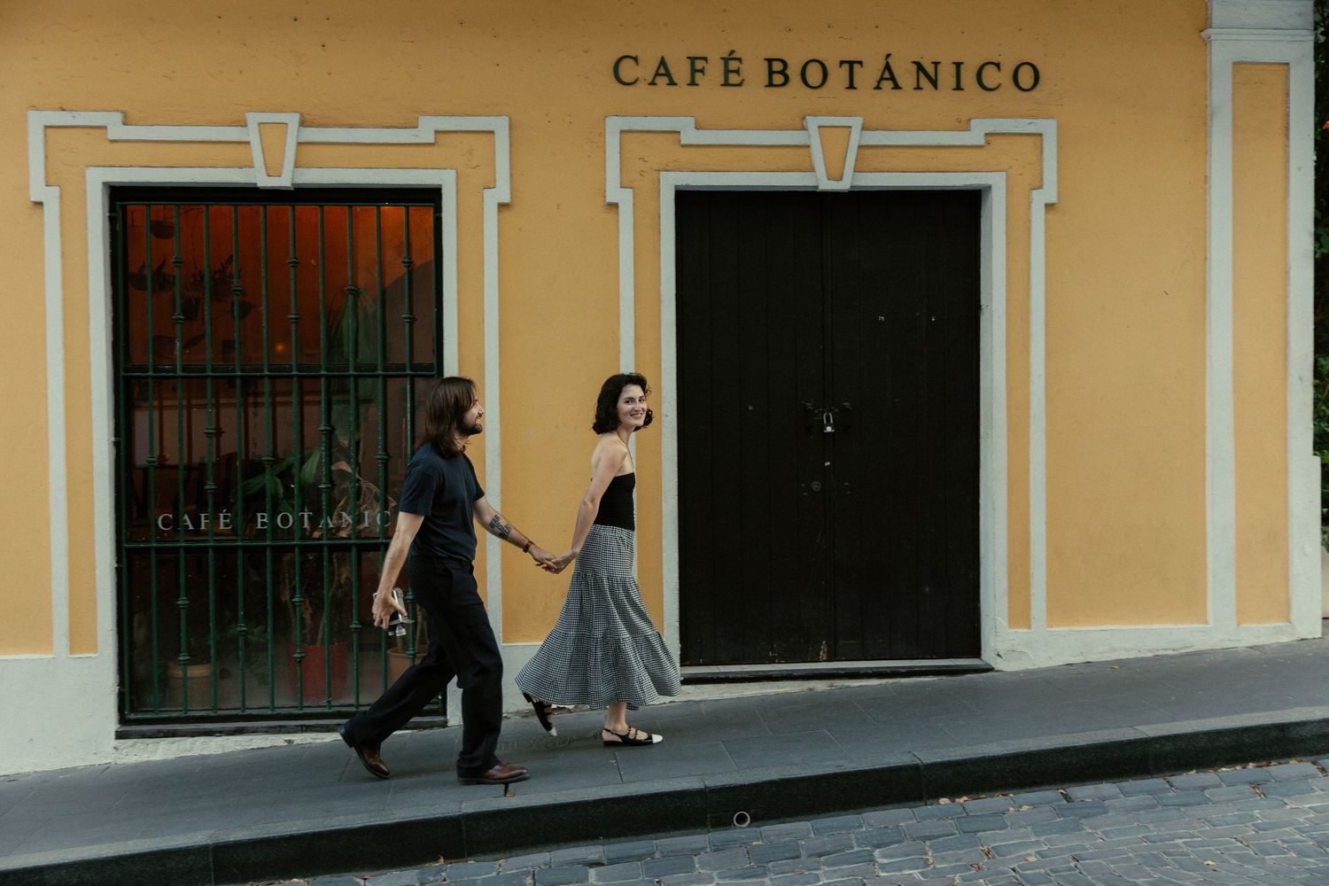 Couple walk hand-in-hand past the yellow facade of Café Botánico on a cobblestone street.