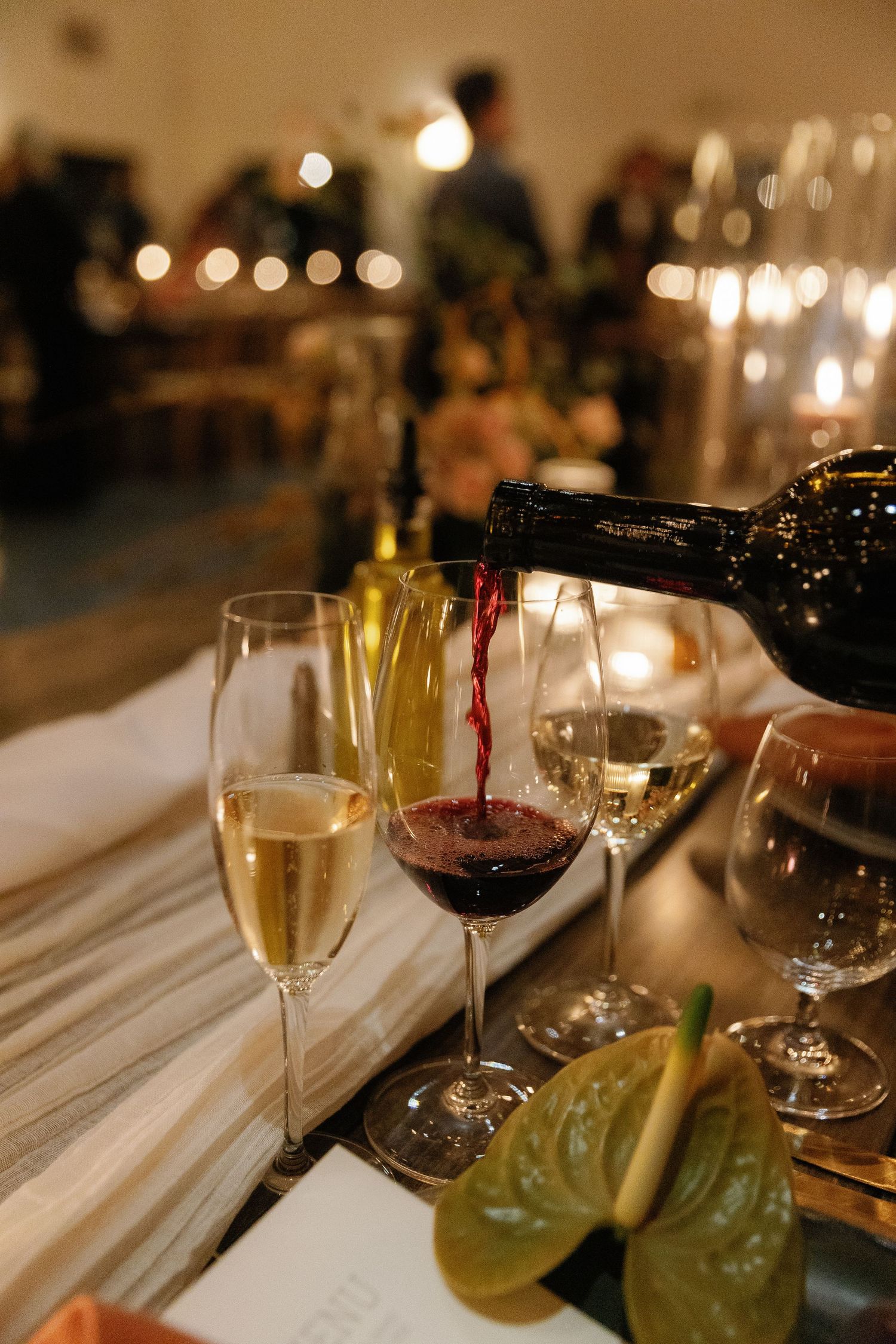 Red wine being poured into glass alongside champagne flute at candlelit wedding reception dinner table.