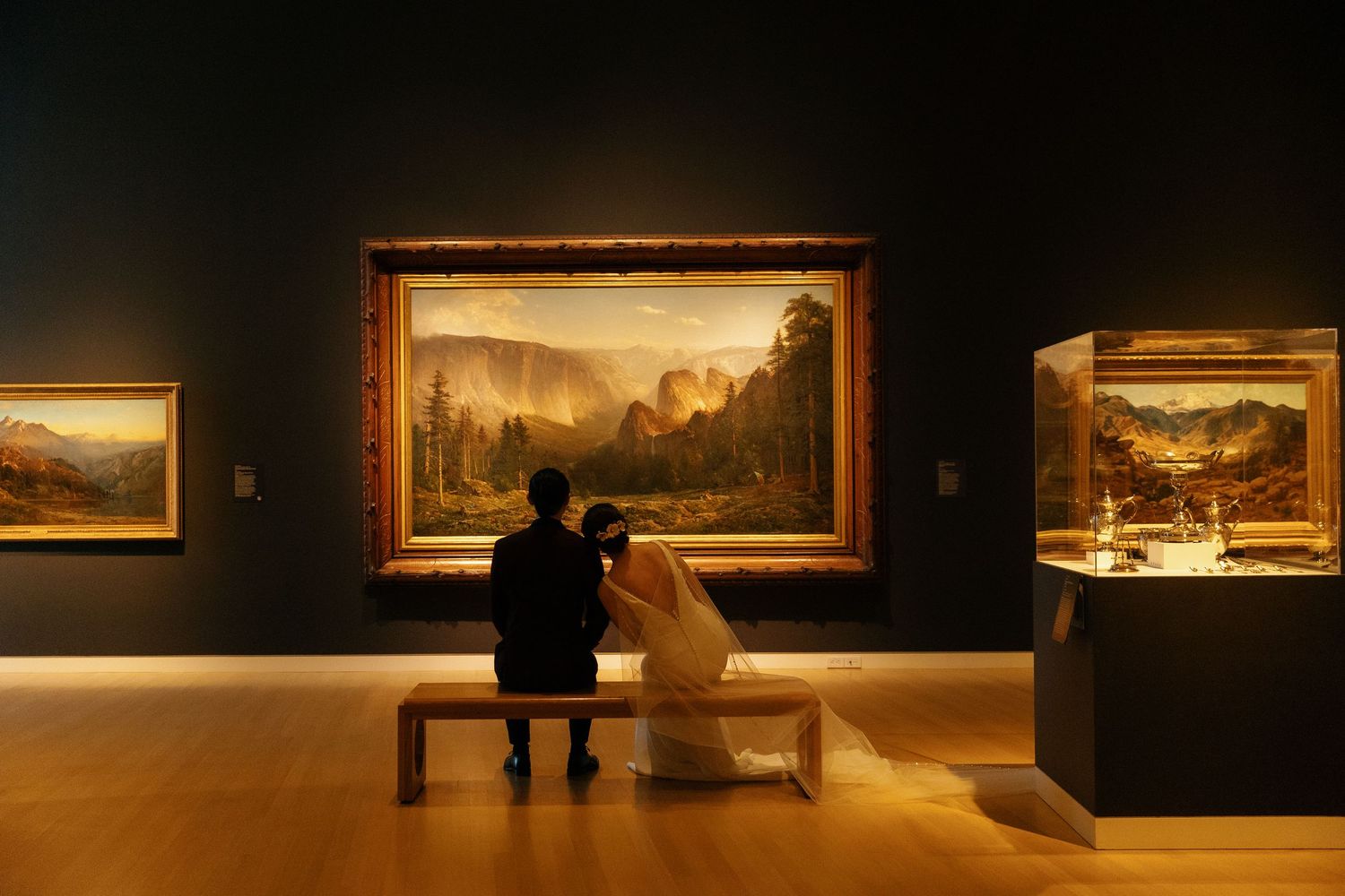 Couple sitting on bench admiring a large landscape painting in a dimly lit art museum gallery.