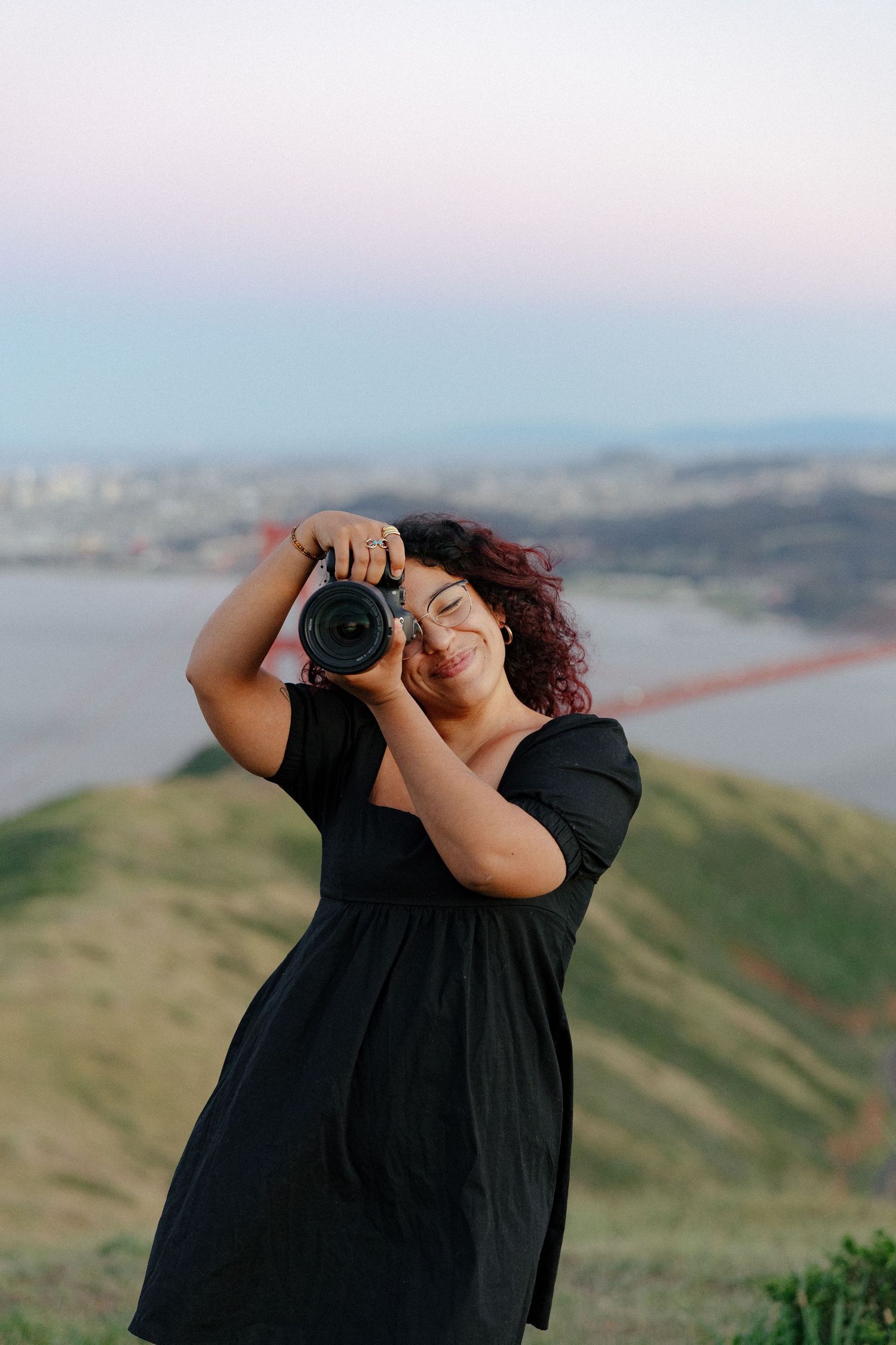 Smiling woman with curly hair holding DSLR camera outdoors with Golden Gate Bridge in background.