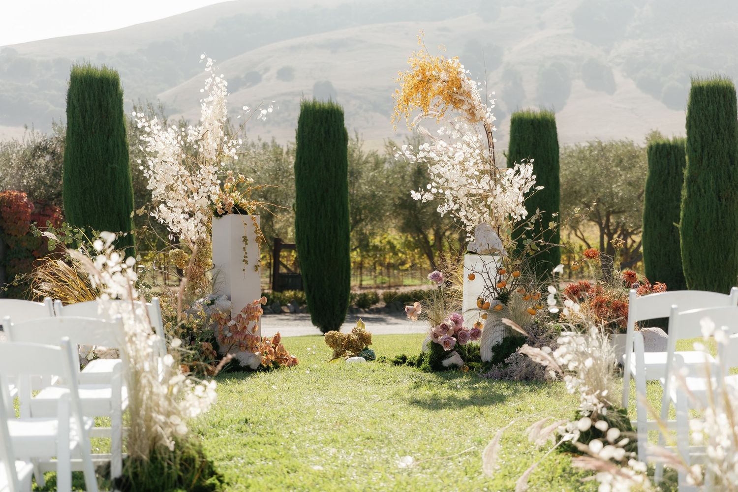 Outdoor wedding ceremony setup with floral arches, white chairs, and scenic vineyard mountain backdrop.