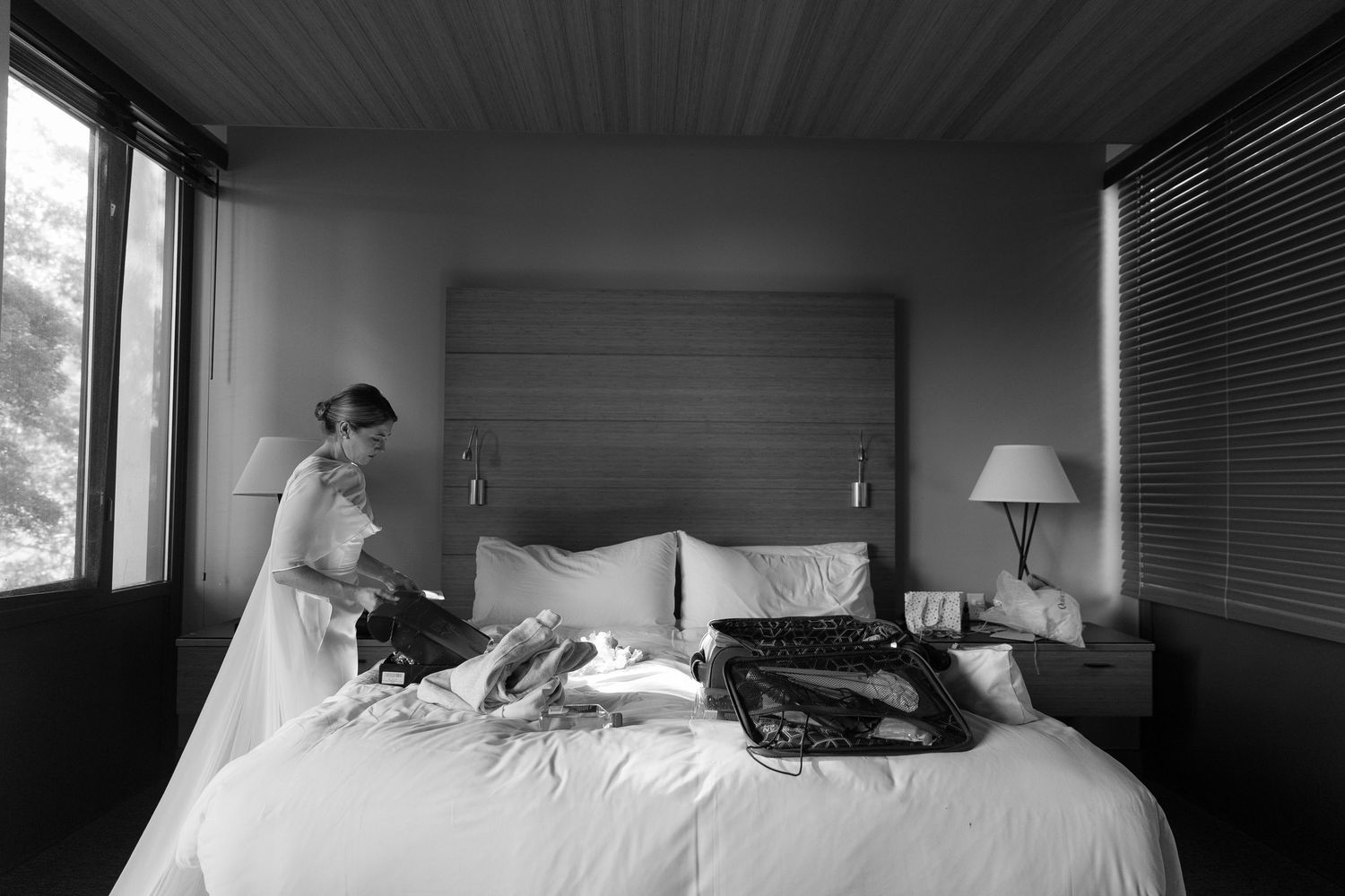 Bride in wedding dress preparing in hotel room with luggage on bed, black and white wedding photography.
