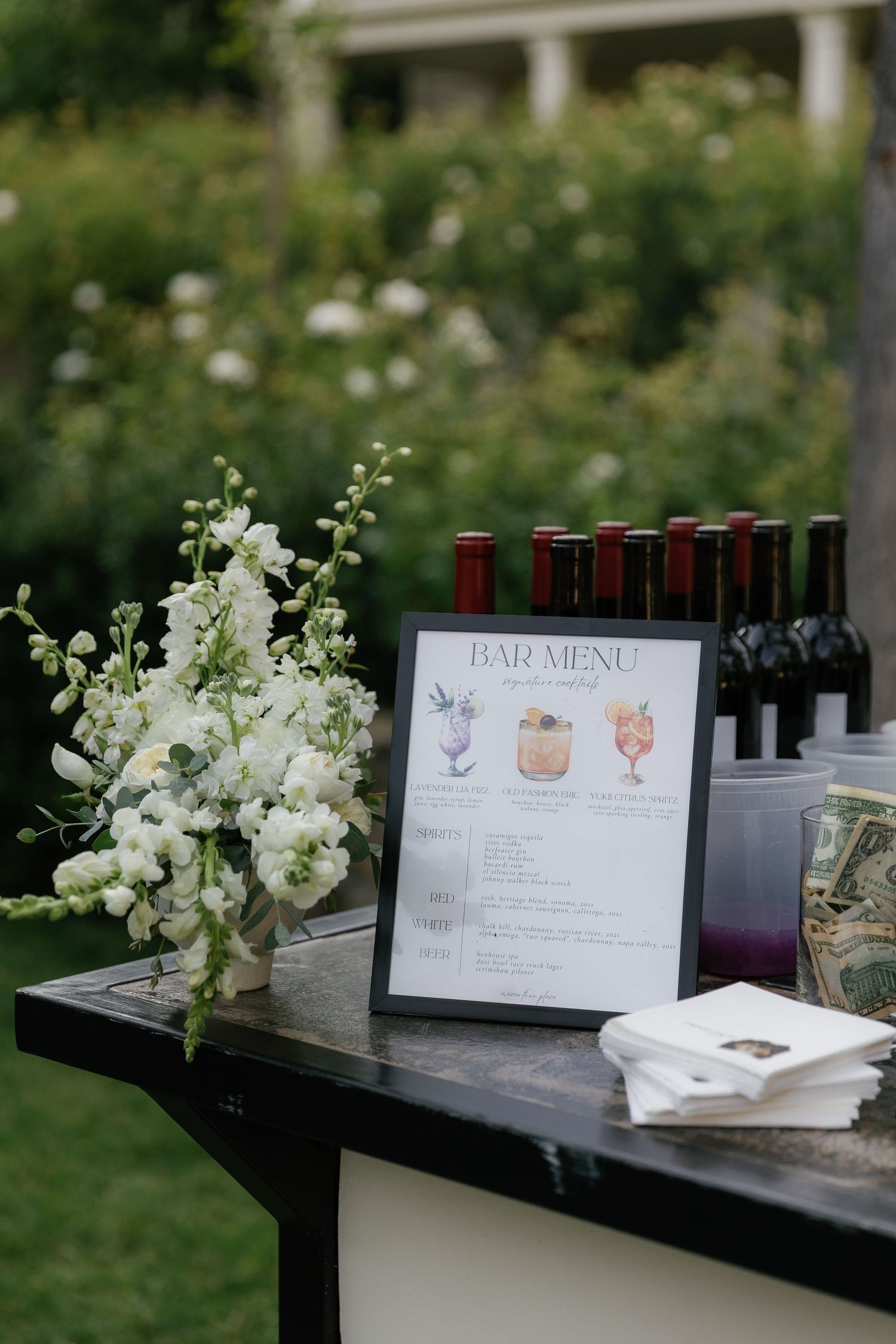 Elegant outdoor bar menu display with white floral arrangement, framed cocktail menu, and wine bottles at a wedding reception.