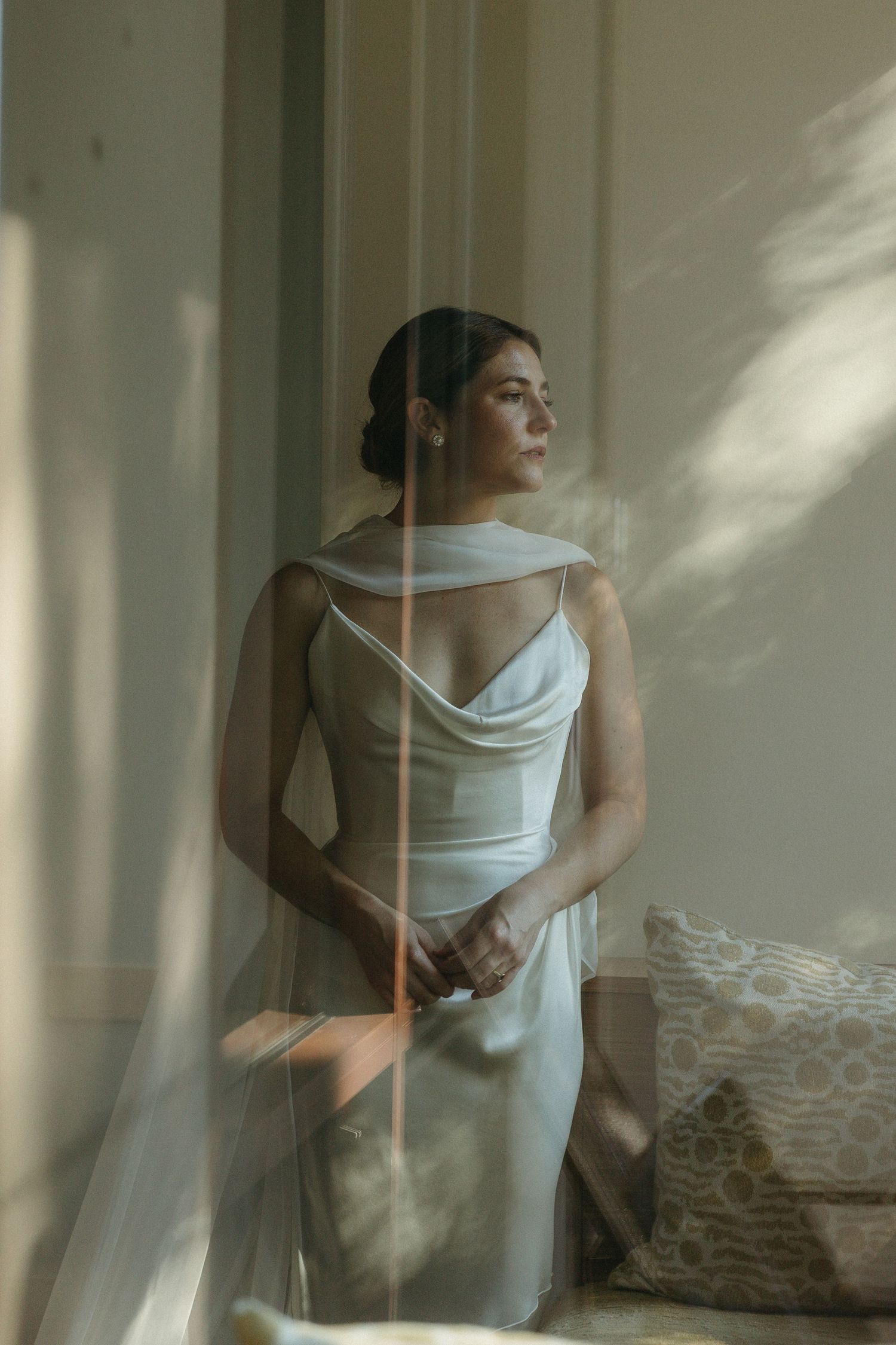 Bride in elegant cowl-neck silk gown with veil standing by sunlit window with warm dappled light.