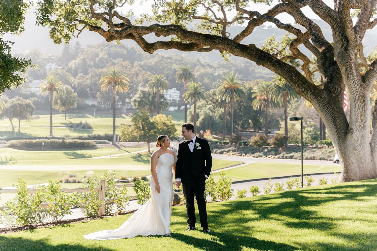 Bride and groom share romantic moment under sprawling oak tree with scenic golf course and palms behind.