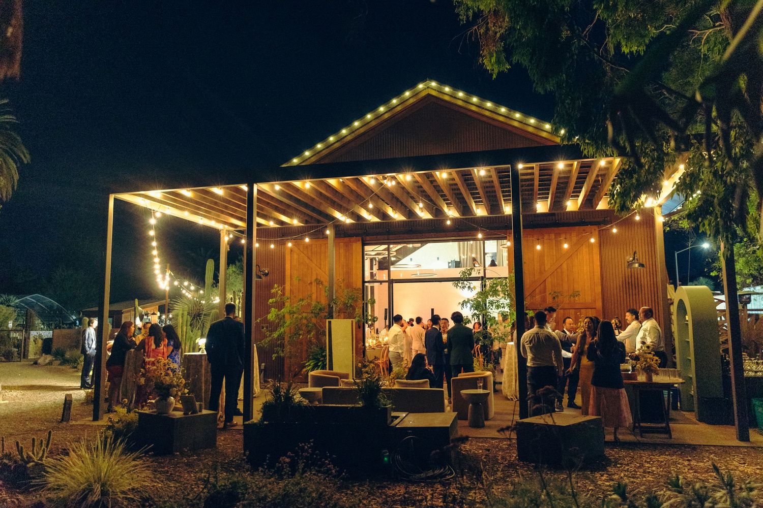 Outdoor evening event venue with string lights illuminating a wooden pavilion surrounded by guests and tropical landscaping.