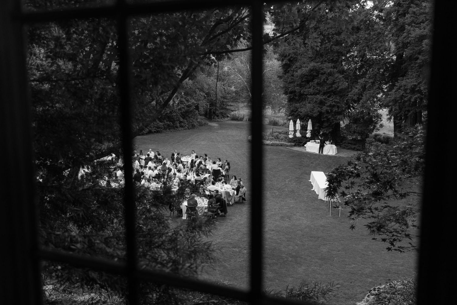 Black and white view through window of outdoor garden wedding ceremony with guests seated on lawn.