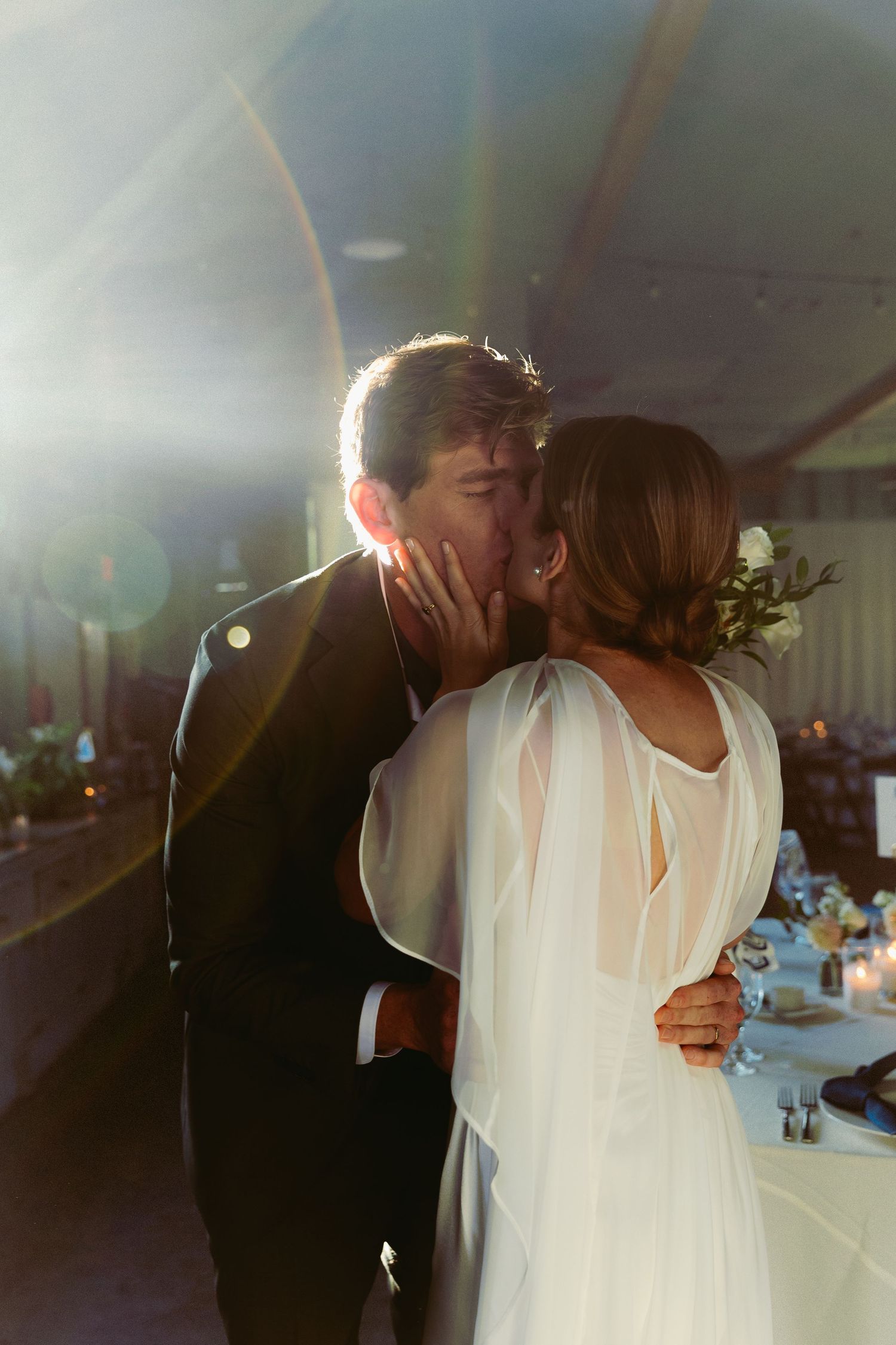 A bride and groom share their first kiss at the reception, bathed in warm lens flare light inside an elegant venue.