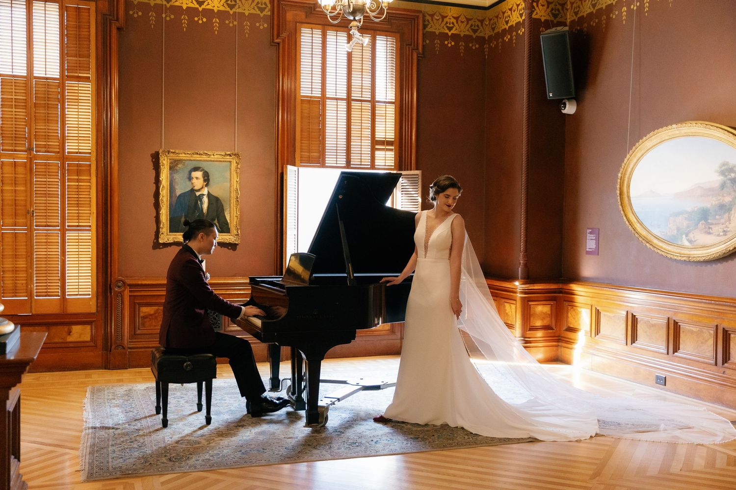 Bride in white gown stands by a grand piano as groom plays in an elegant wood-paneled room.