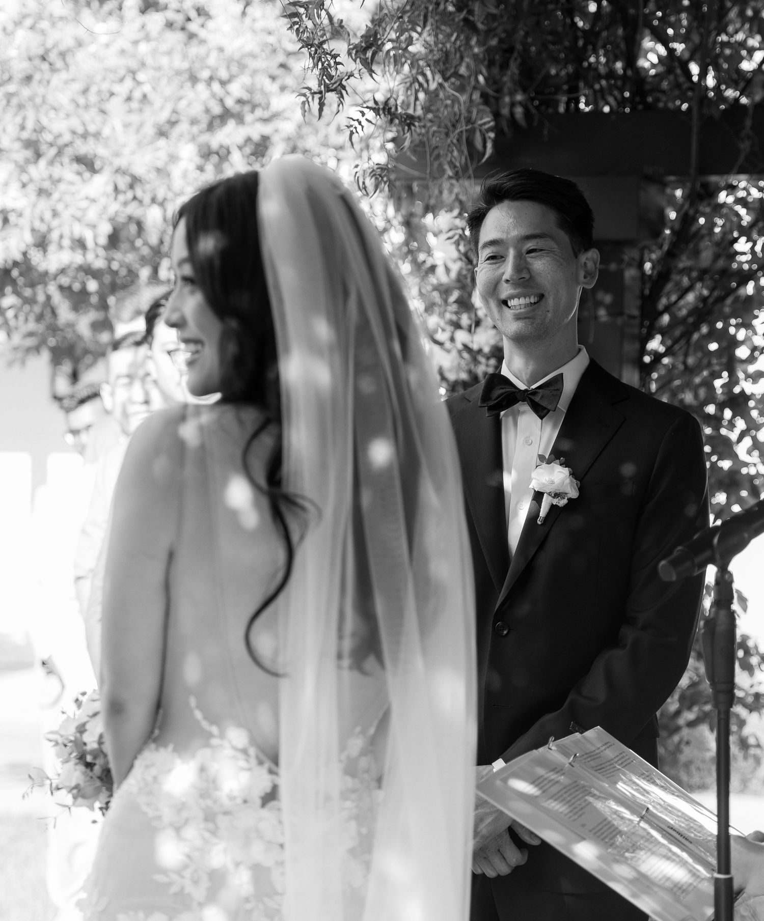 Black and white photo of smiling groom watching bride during outdoor wedding ceremony.