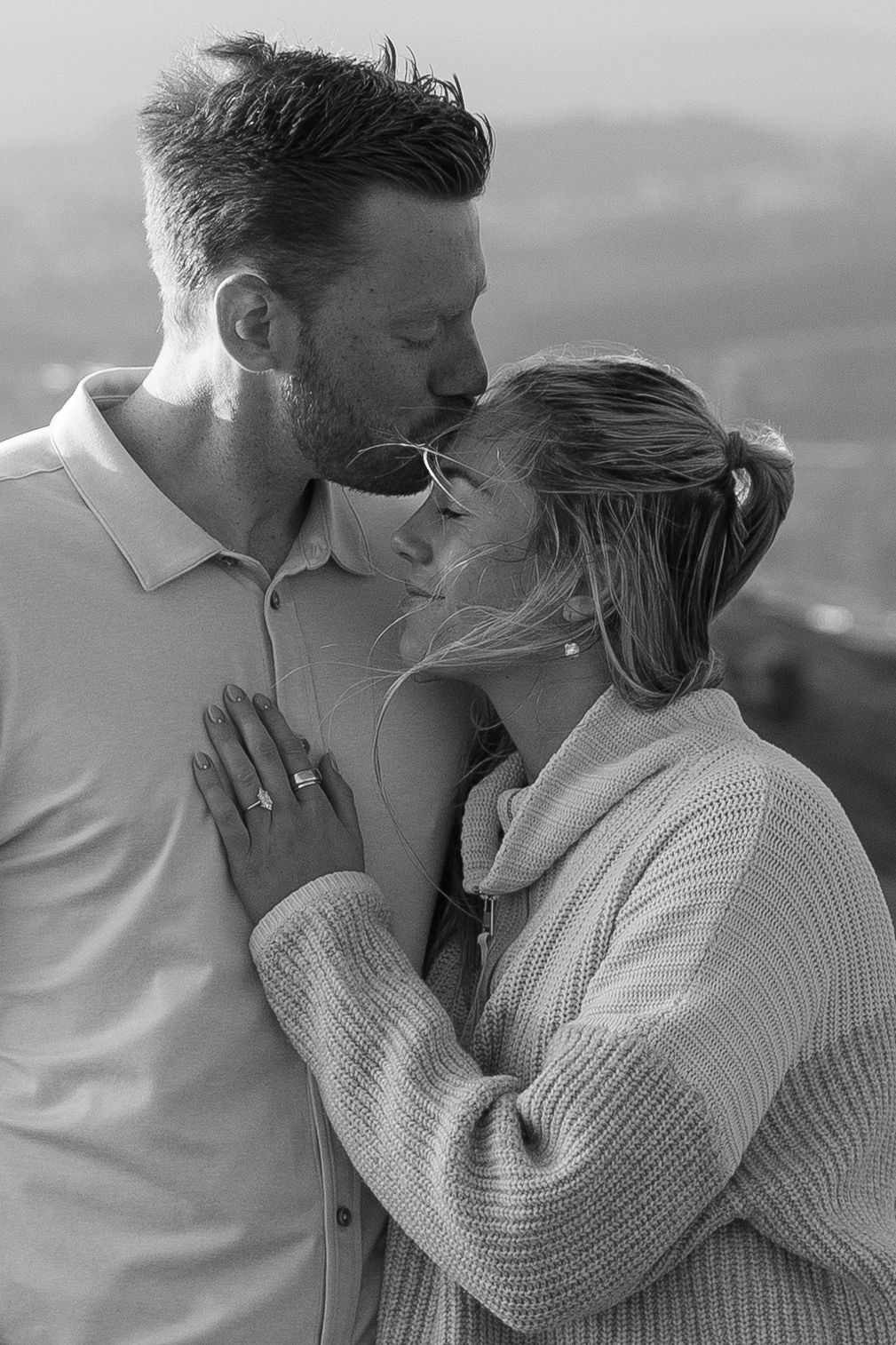 Black and white photo of a couple sharing a romantic moment, woman displaying engagement ring, outdoors with scenic backdrop.