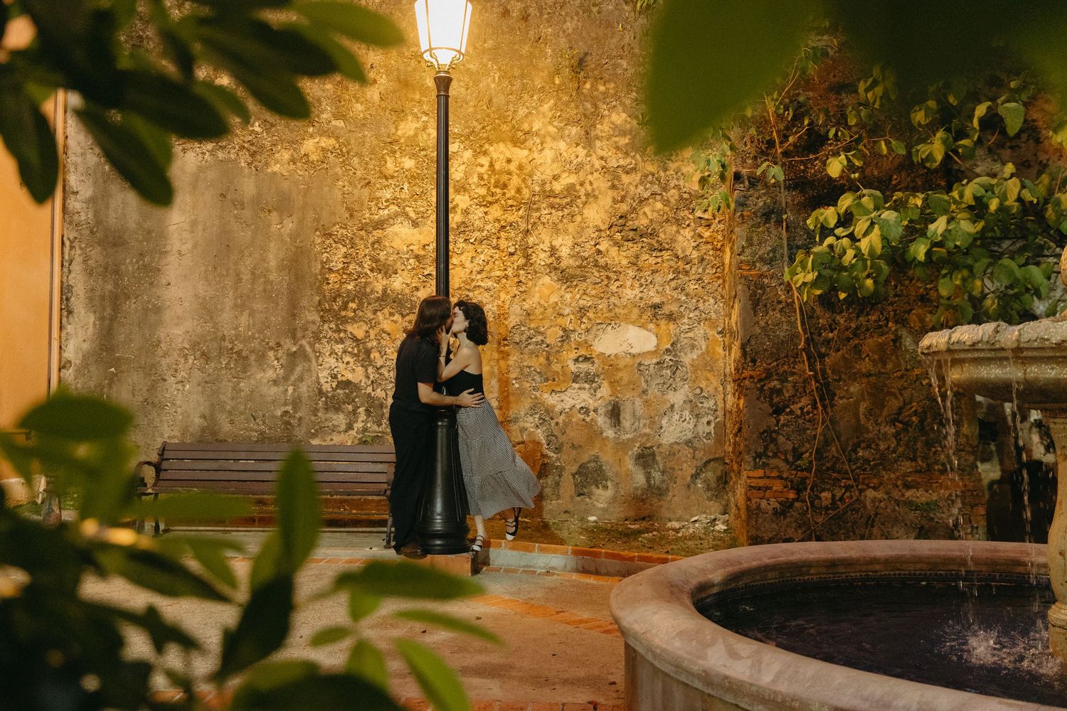 A couple stands embracing under a glowing street lamp next to a stone fountain in a romantic garden setting at night.