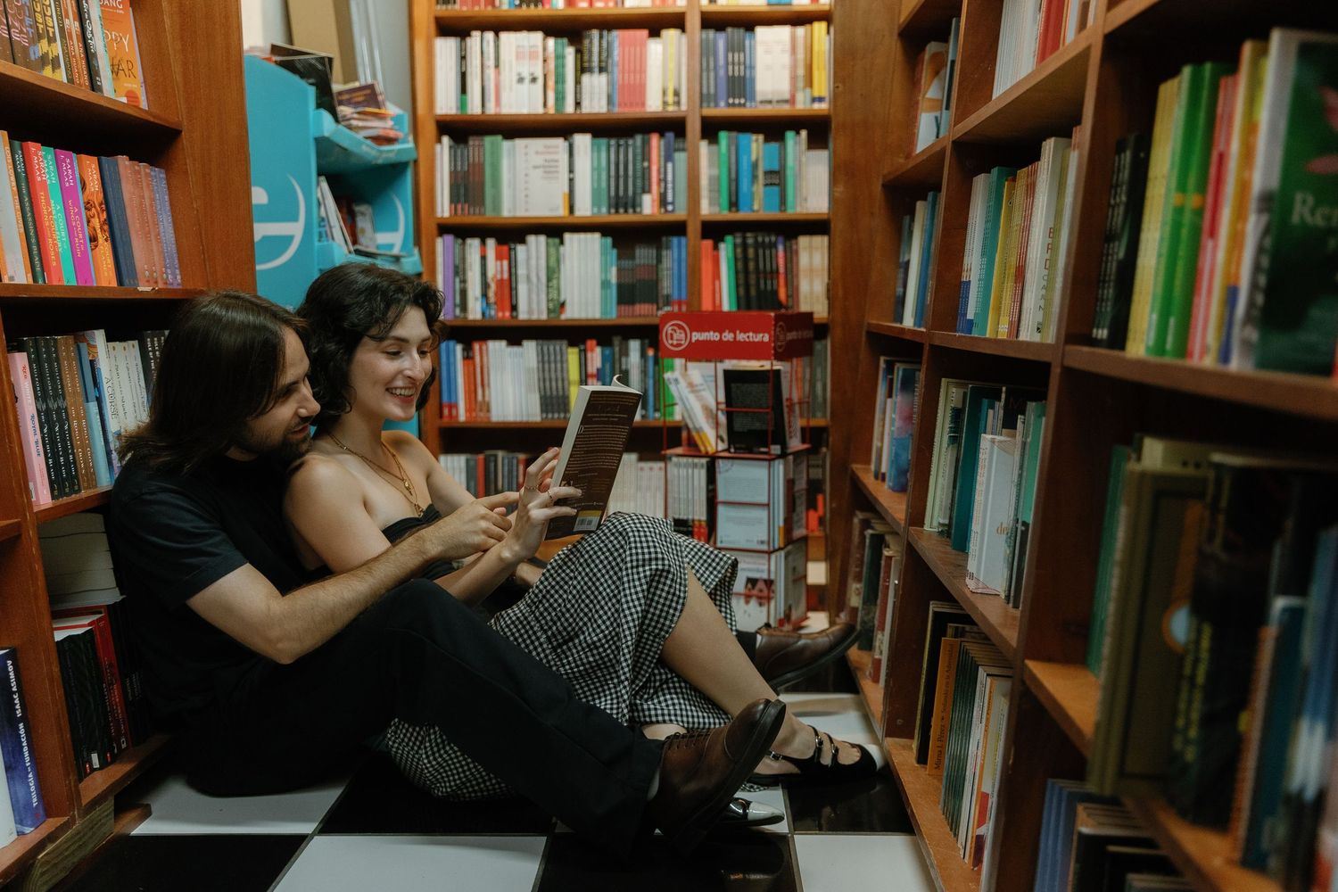 Two people sit together on the floor of a bookstore browsing their phones while surrounded by bookshelves.