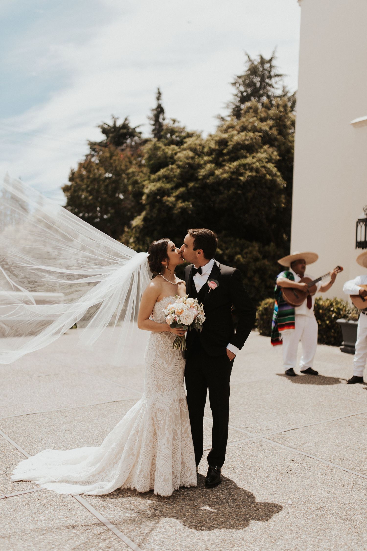 Bride and groom share a kiss outdoors, her veil flowing in the wind, as a mariachi musician performs behind them.