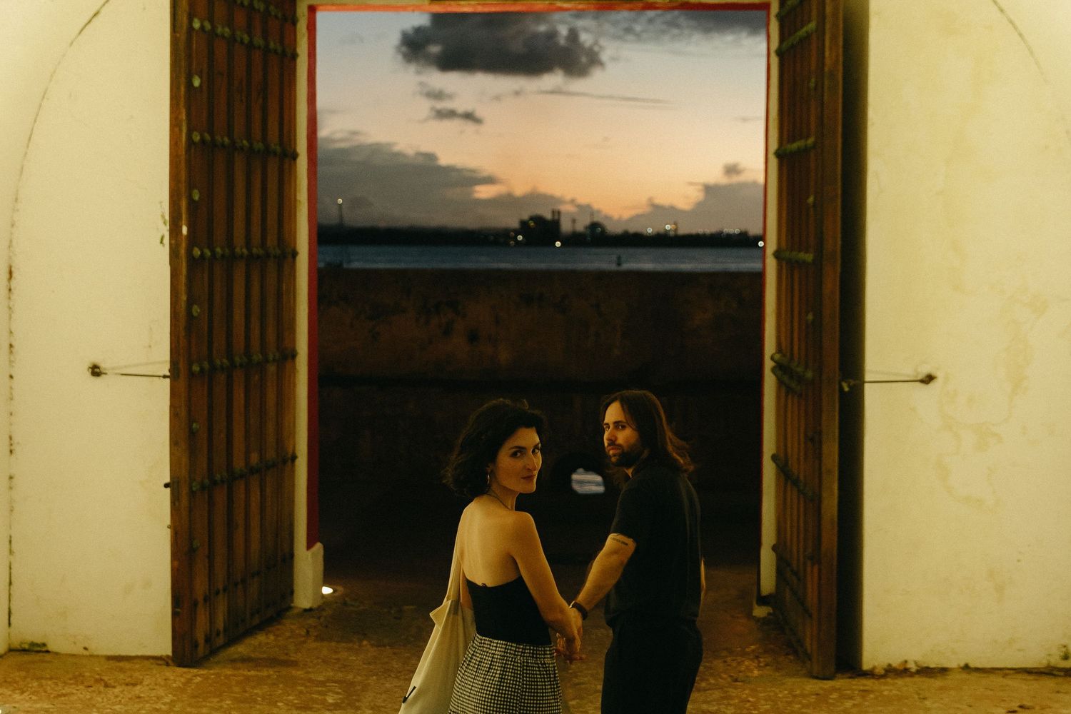 Two people stand in silhouette at a doorway against a sunset sky over a field.