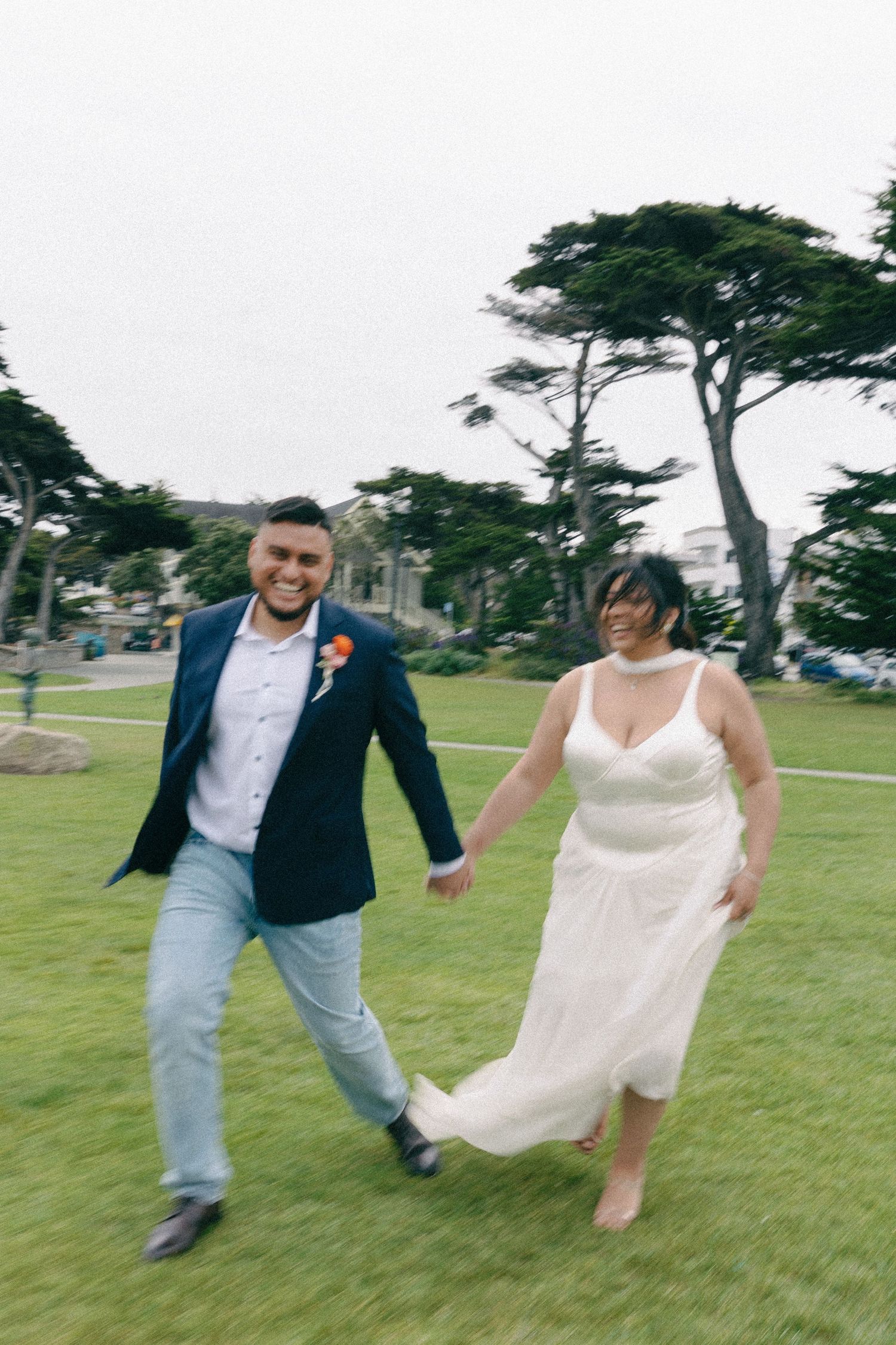 Joyful newlyweds run hand-in-hand on green grass, bride in white dress, groom in navy blazer, outdoors.