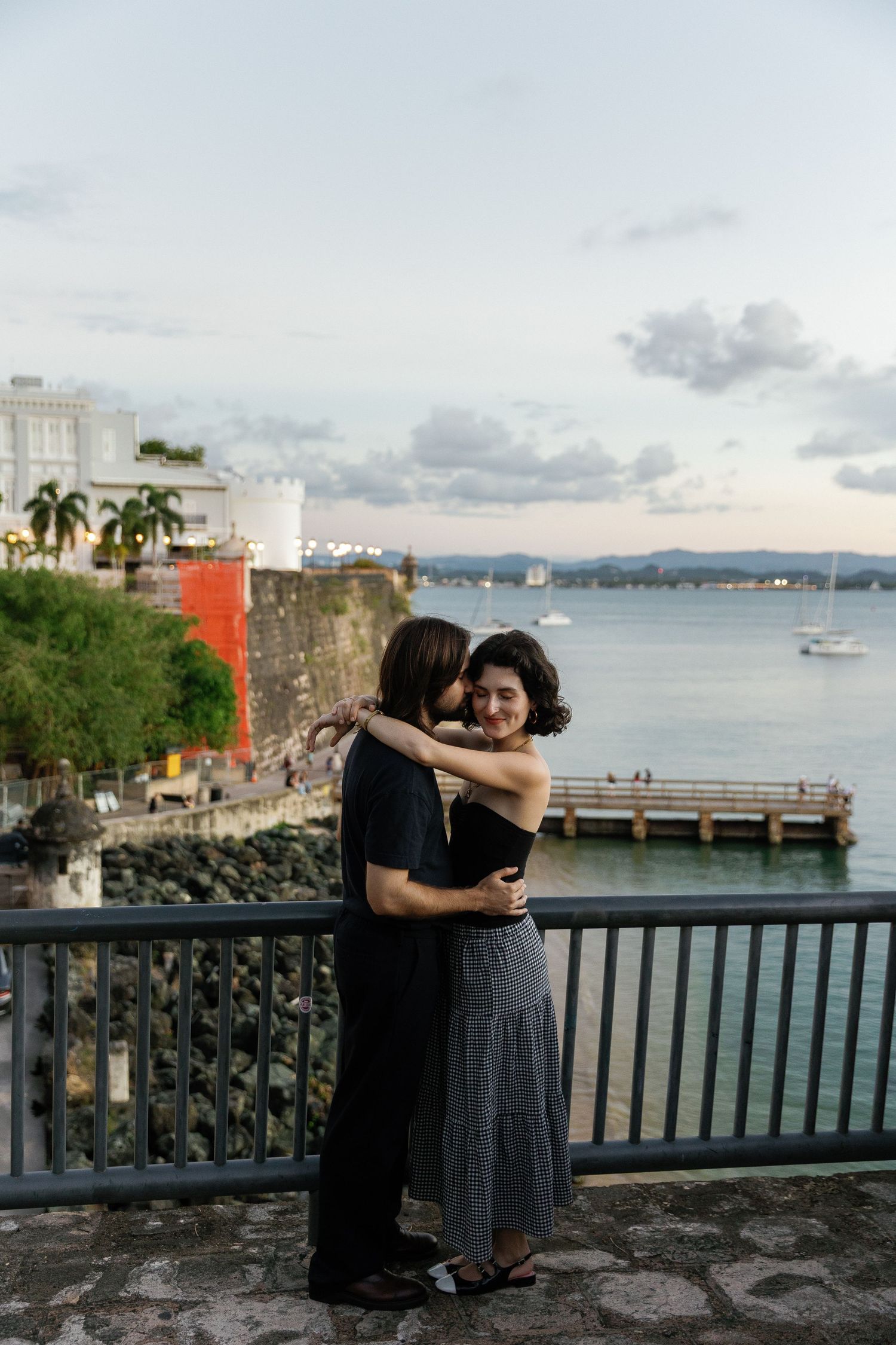 Two figures embrace on a waterfront walkway with historic fortification walls and harbor views in Old San Juan, Puerto Rico.