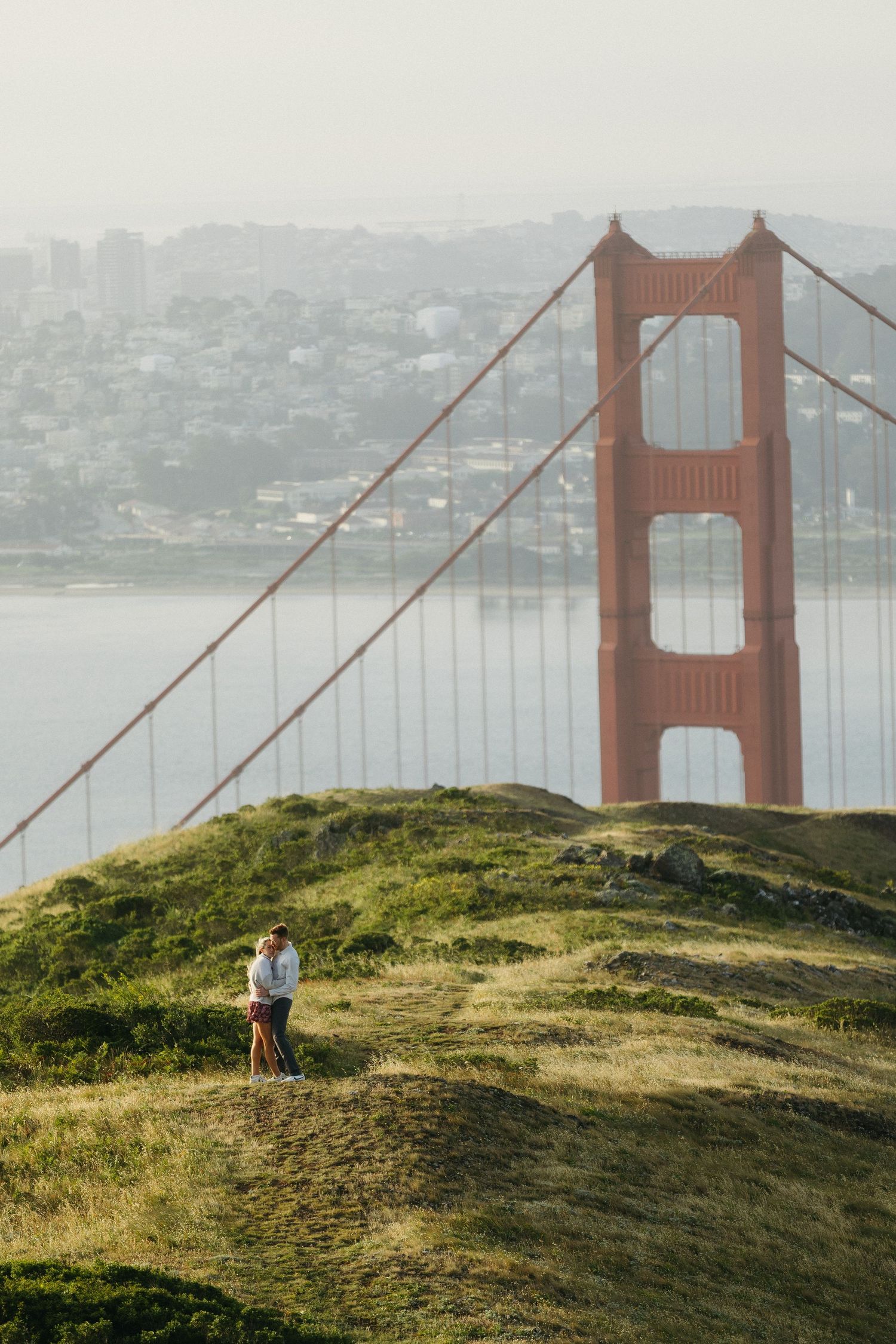 Couple embracing on a grassy hillside with the Golden Gate Bridge towering in the misty background.