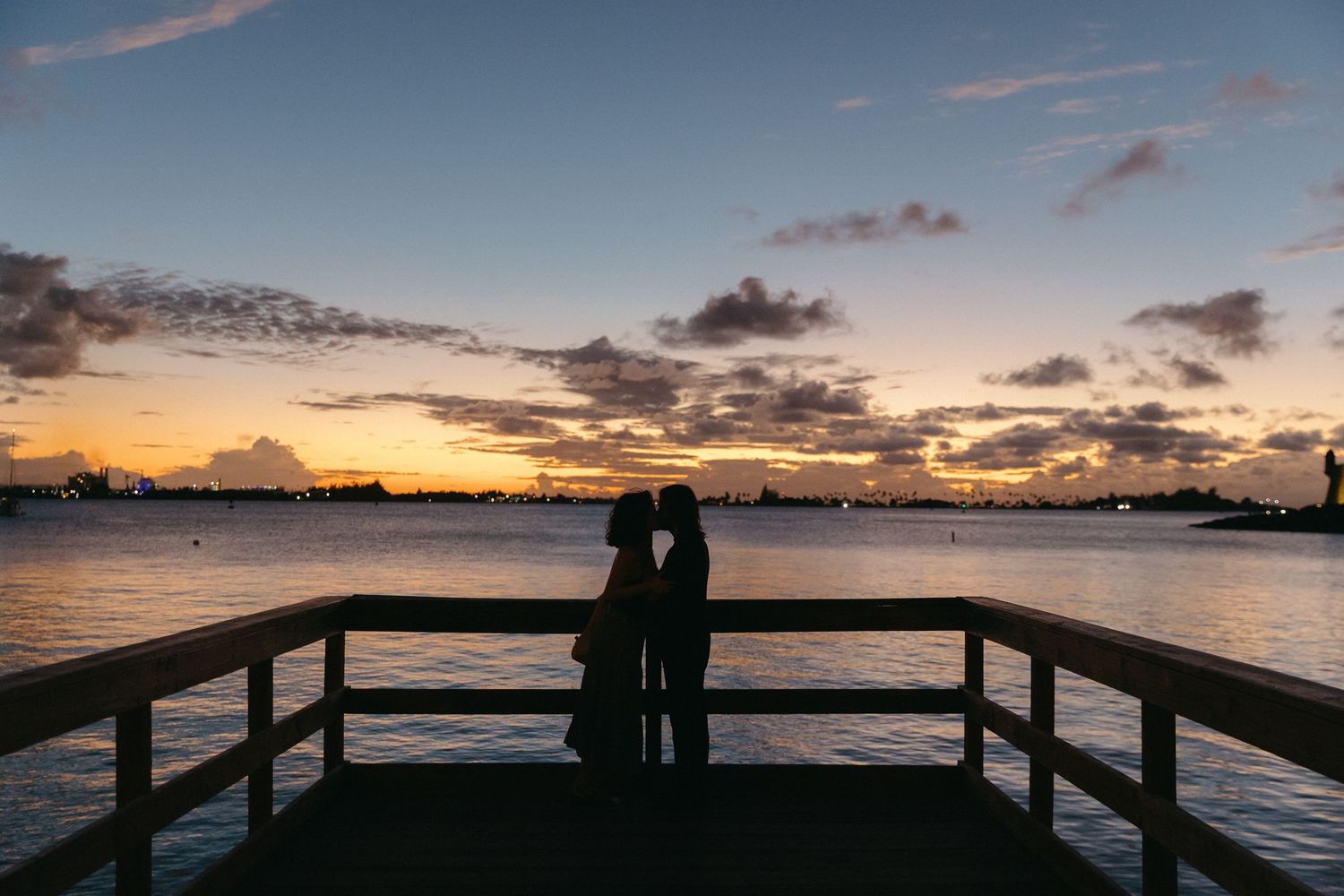 Silhouetted couple standing on wooden pier watching vibrant sunset over calm water with cloudy sky.
